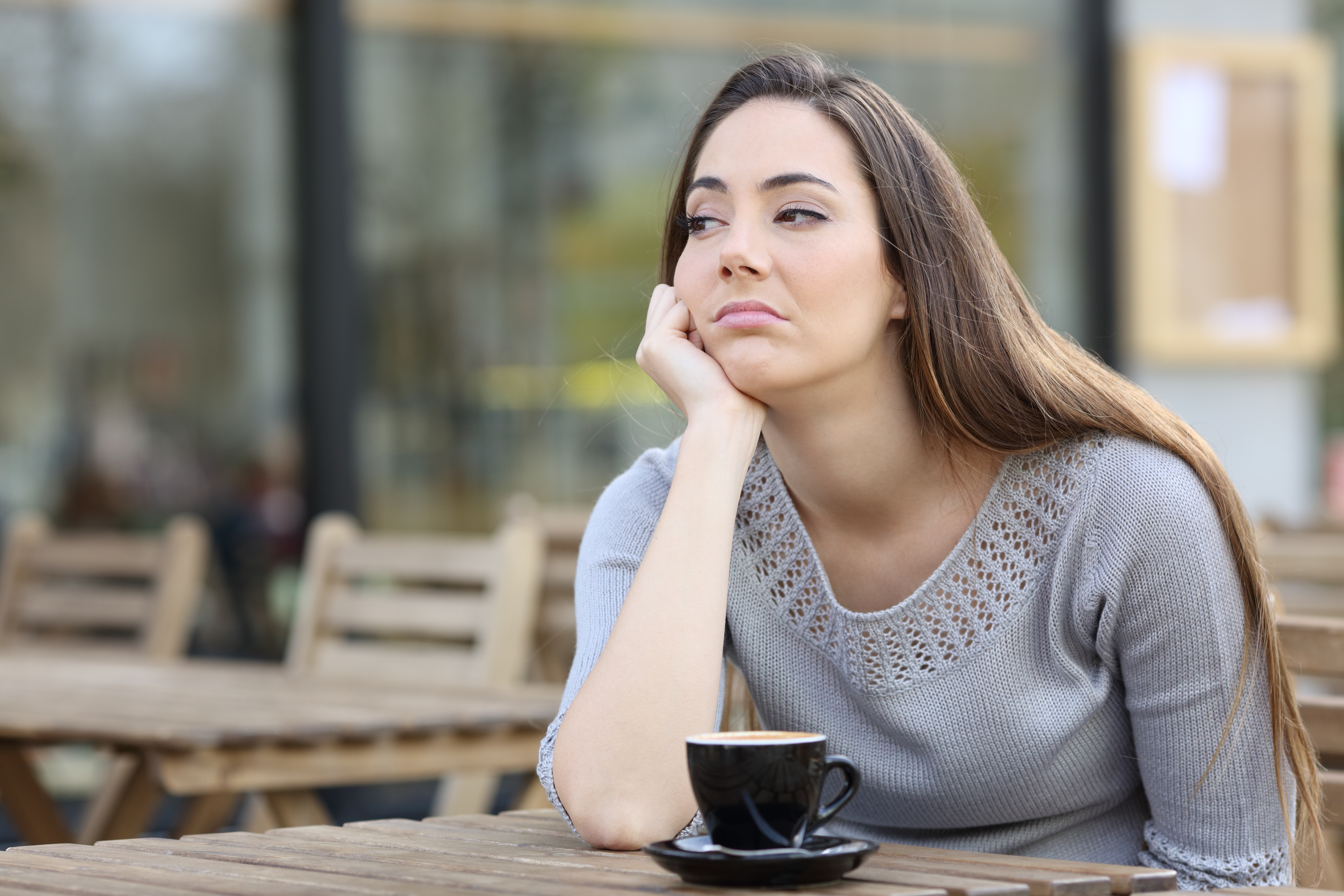 Person sitting at an outdoor cafe, looking pensive with their chin resting on their hand, a cup of coffee in front of them