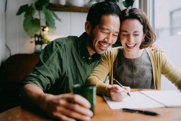 A happy couple sits at a table, smiling and working together on a notebook. One holds a cup, while the other writes. Plants are visible in the background