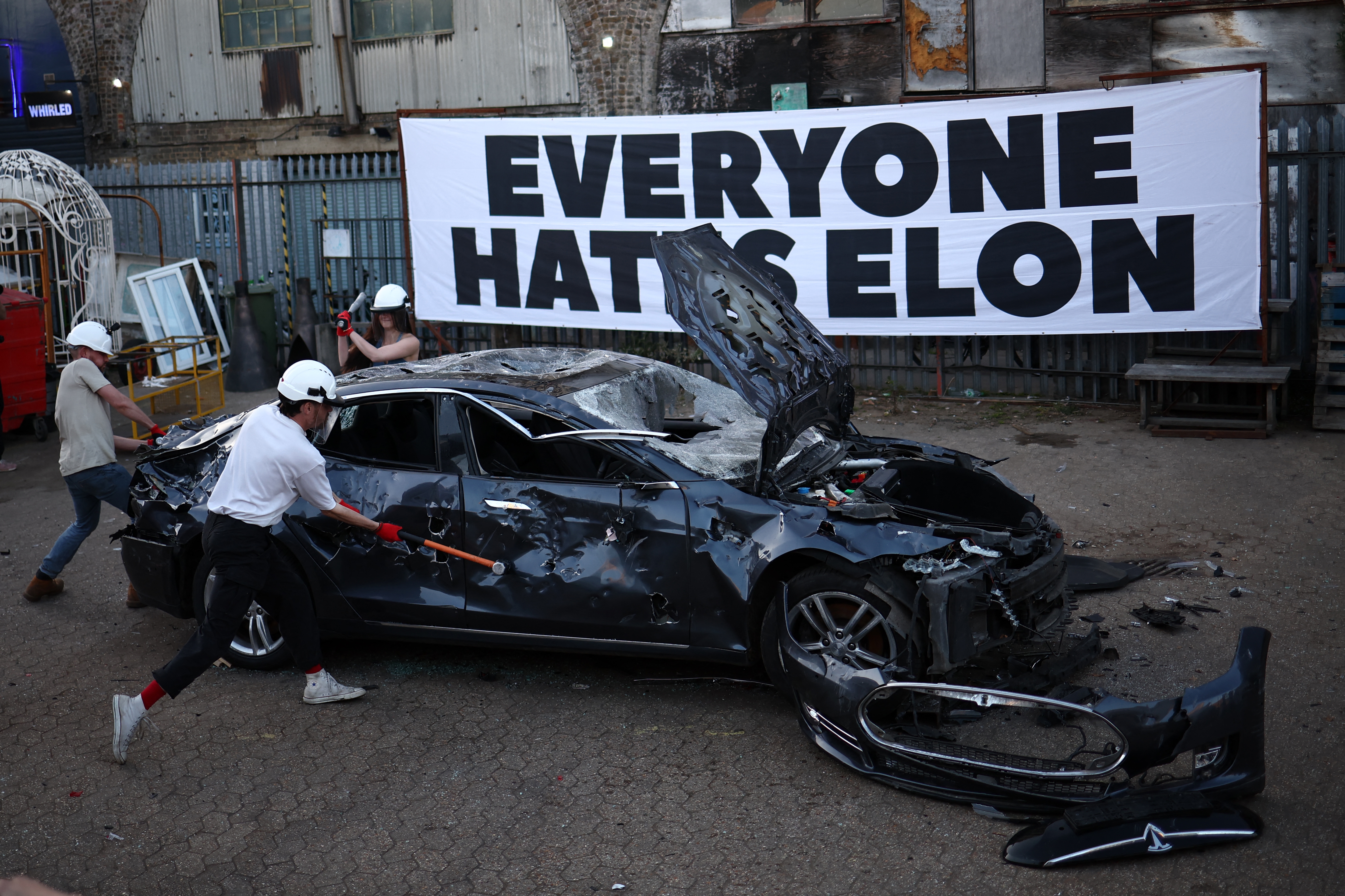 People smash a wrecked Tesla car with hammers in front of a large sign reading "Everyone Hates Elon"