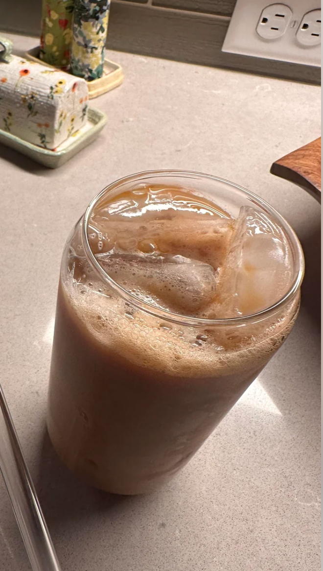 Iced coffee with foam in a glass on a kitchen counter near floral items and an electrical outlet