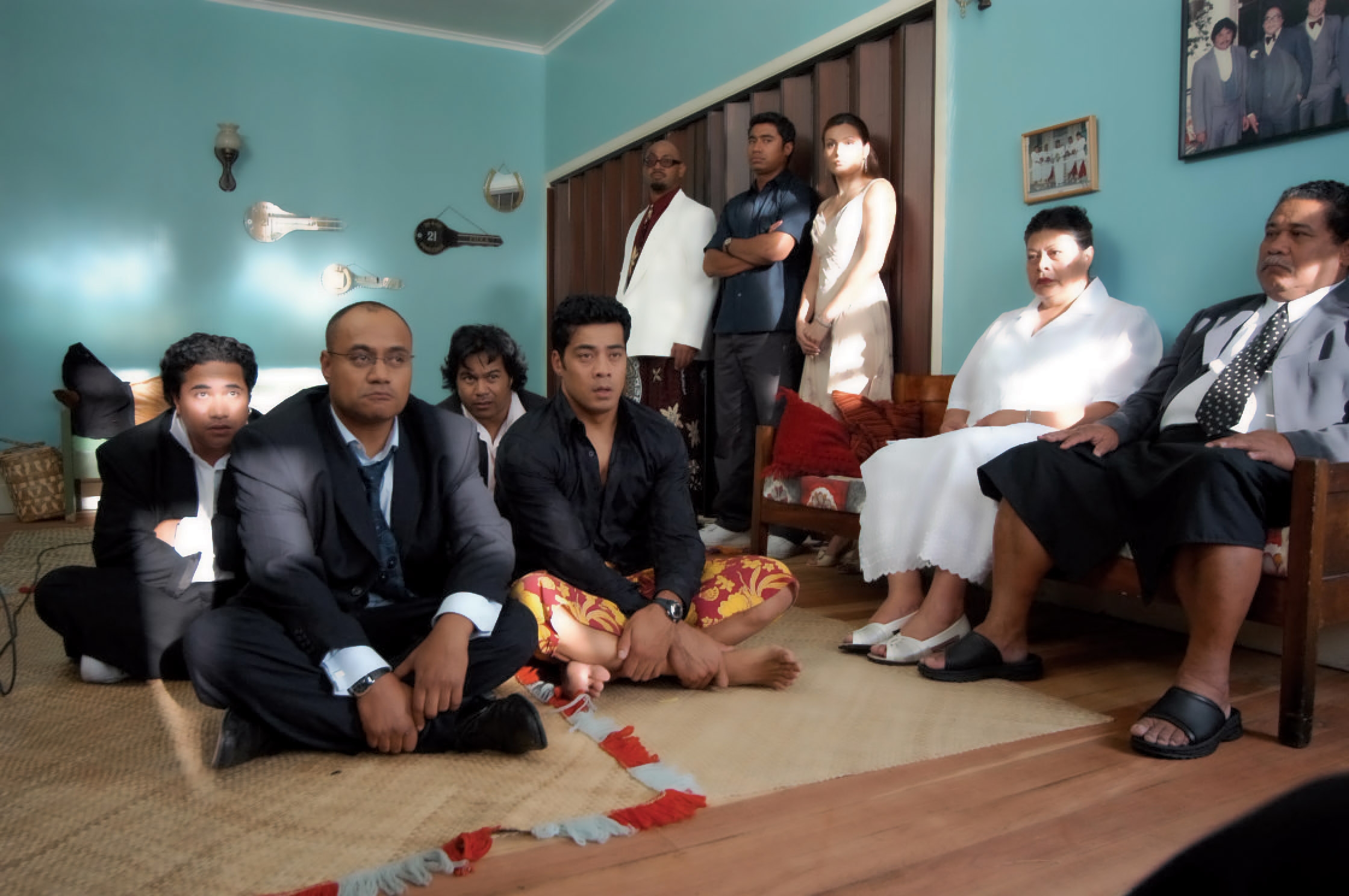 A diverse group of people gather in a living room, some seated on the floor in traditional attire, others on sofas. They appear solemnly attentive