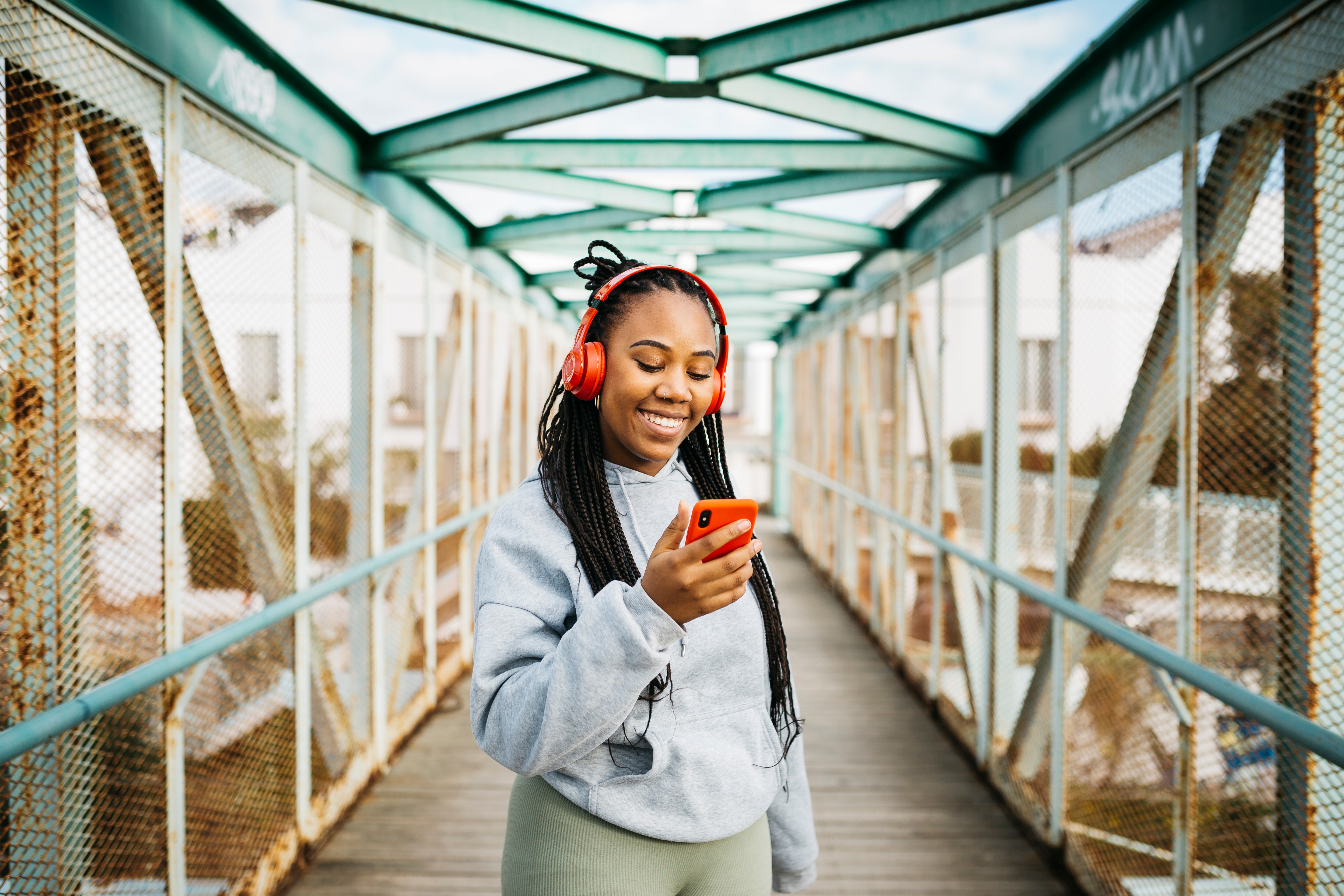 Person with headphones smiling and looking at smartphone on a bridge, wearing a hoodie and leggings, enjoying a moment while walking