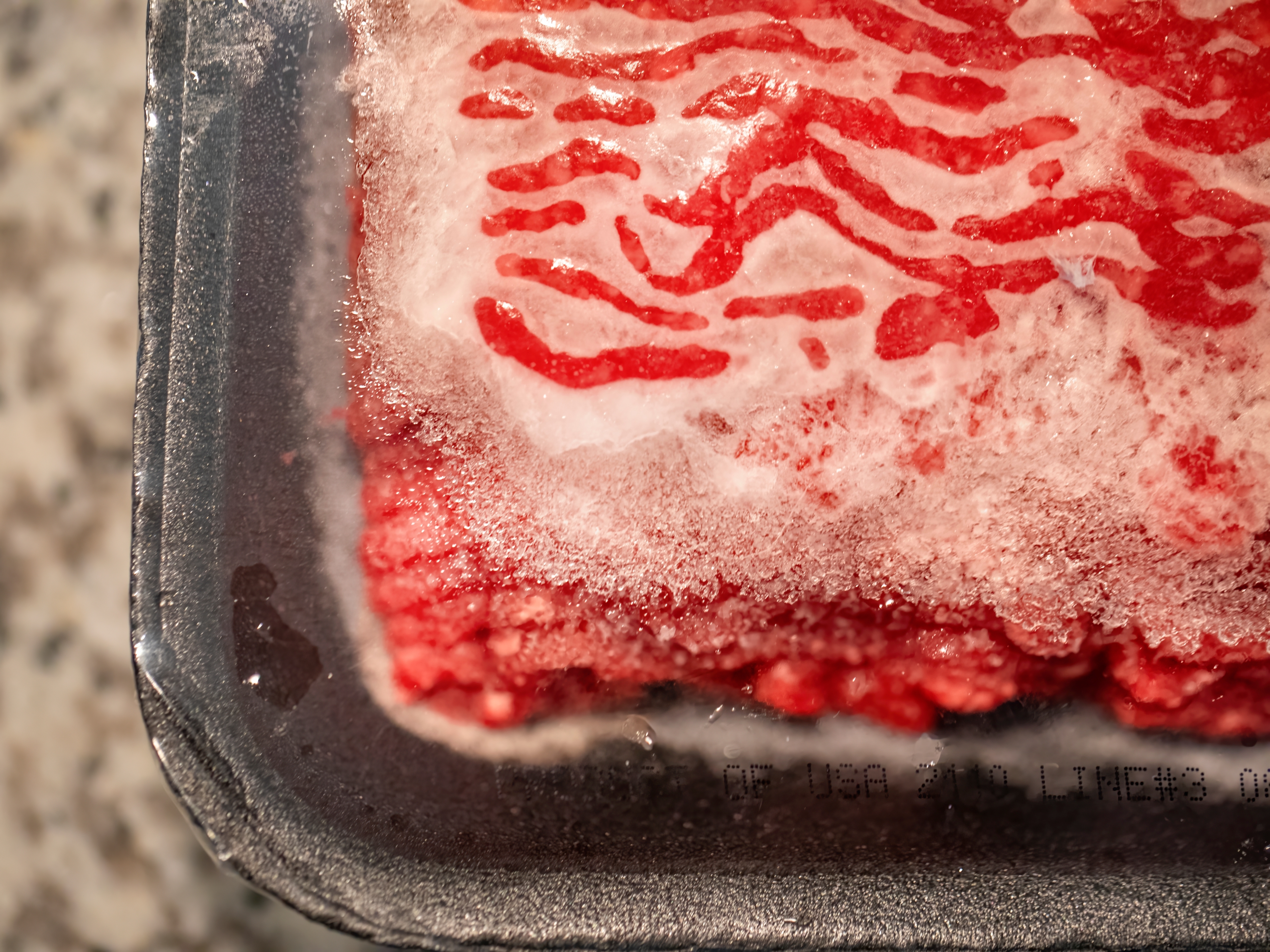Frozen ground beef with icy coating in a metal tray on a speckled countertop