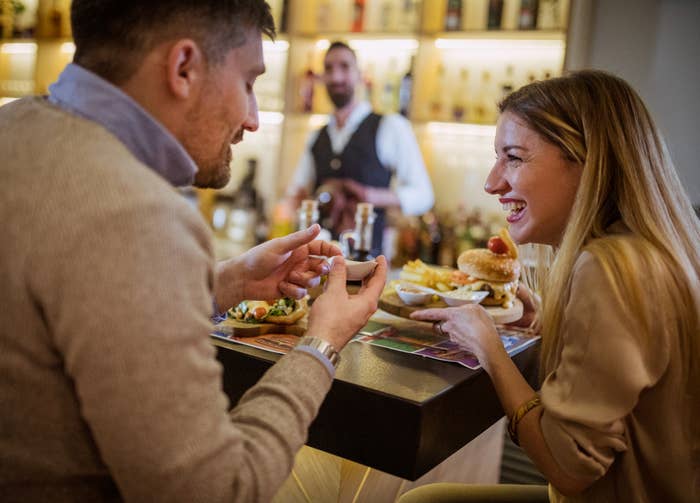 A couple shares a laugh over food and drinks at a bar, with a bartender in the background