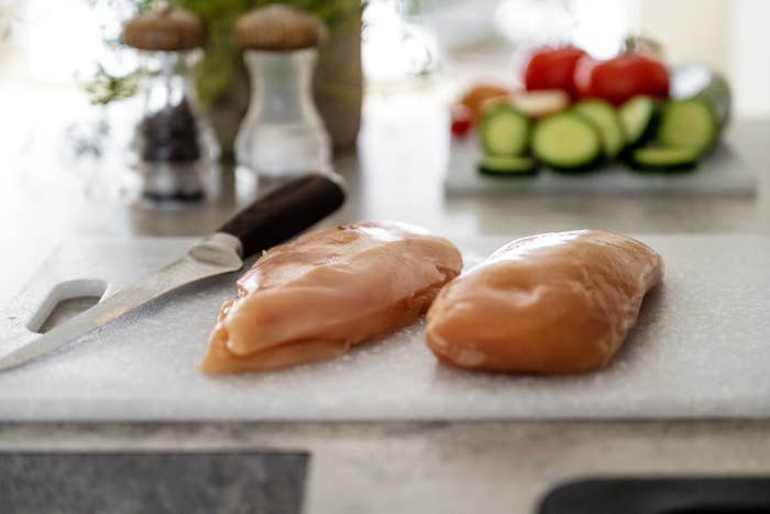 Two raw chicken breasts on a cutting board with a knife nearby; vegetables are blurred in the background