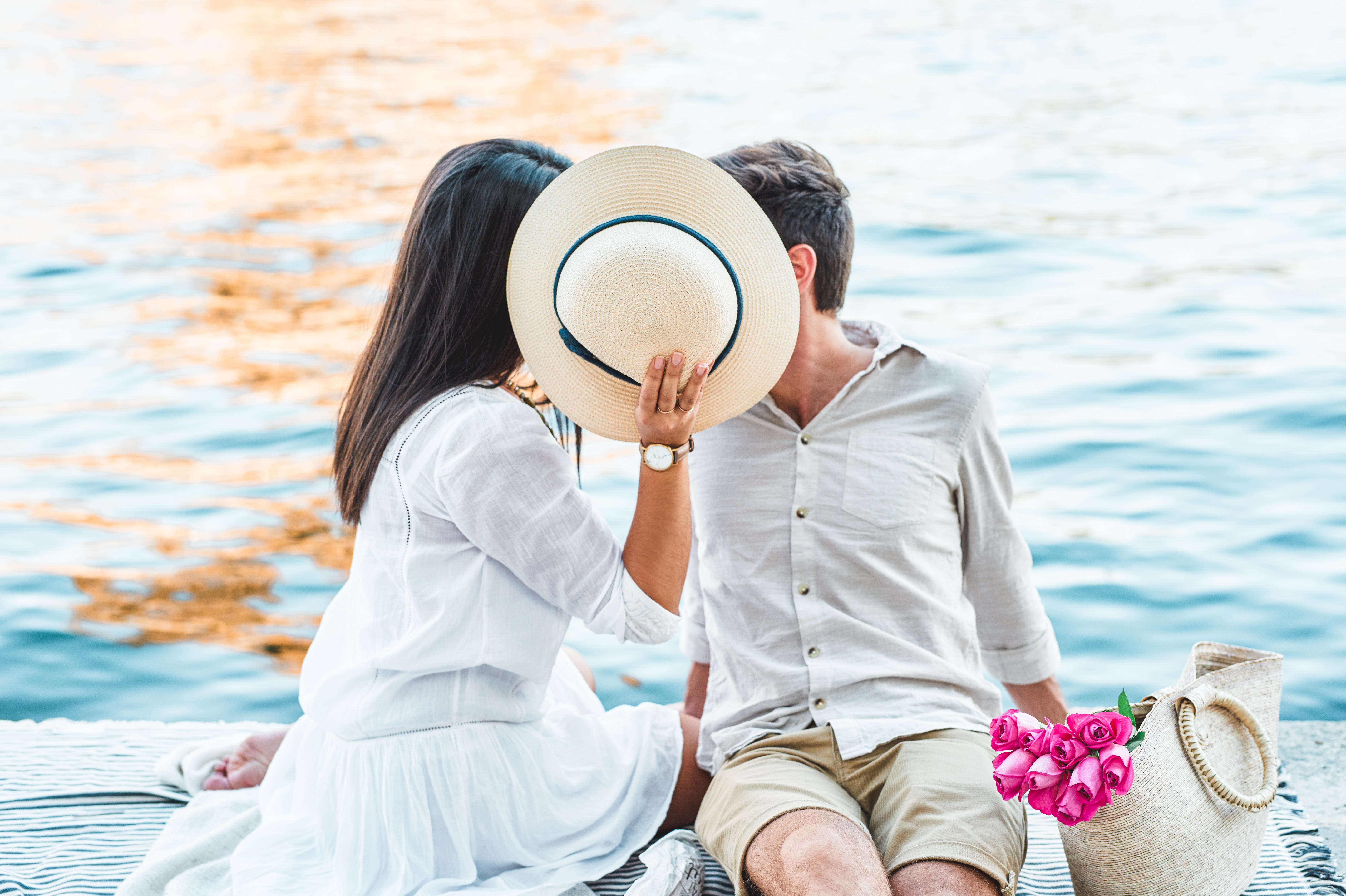 A couple sits by the water, sharing a kiss hidden behind a sun hat. A nearby basket holds pink flowers