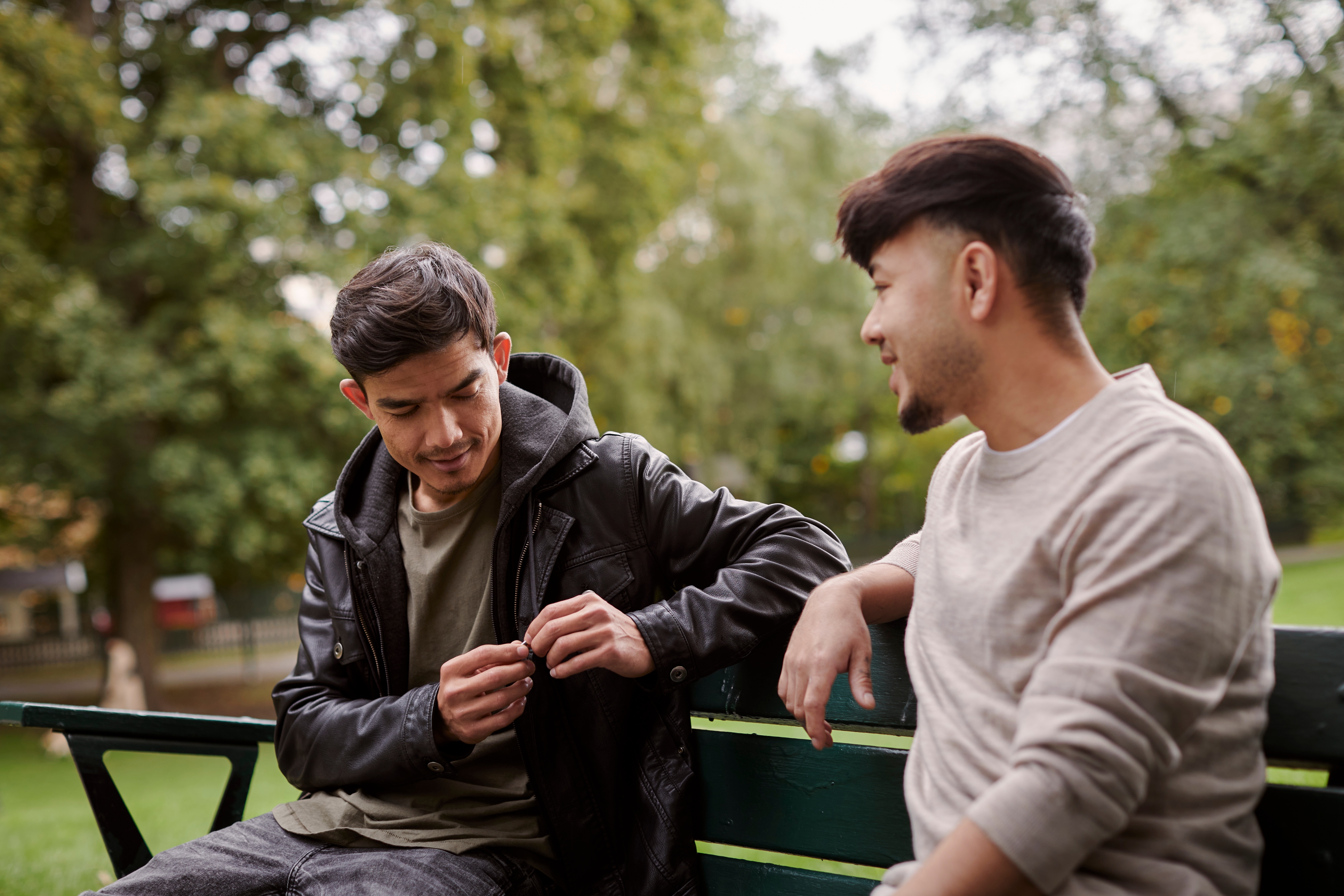 Two people sitting on a park bench, engaged in conversation, with one person adjusting their jacket