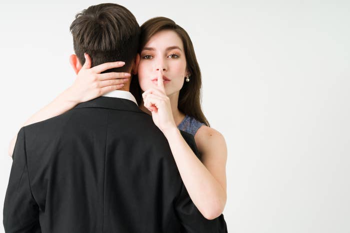 Woman in a sleeveless dress holds a man in a suit from behind, making a shushing gesture with her finger to her lips