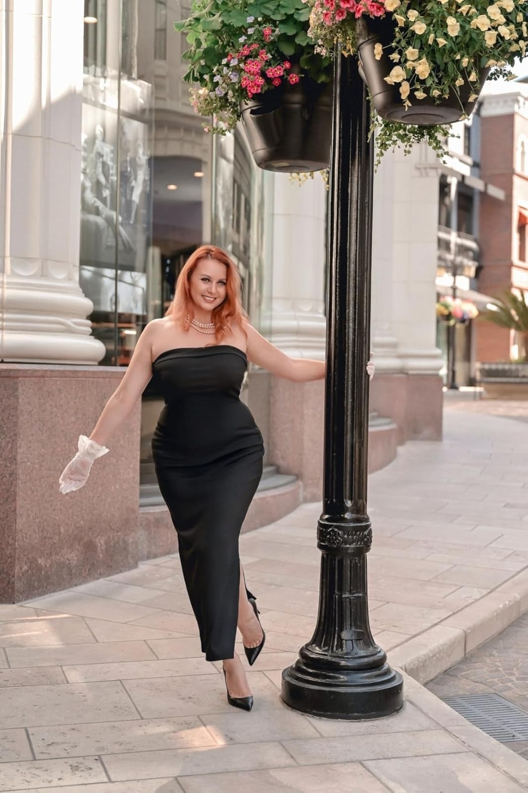 Person successful  a strapless achromatic  evening gown and gloves stands outdoors by a lamppost, showcasing an elegant buying  outfit