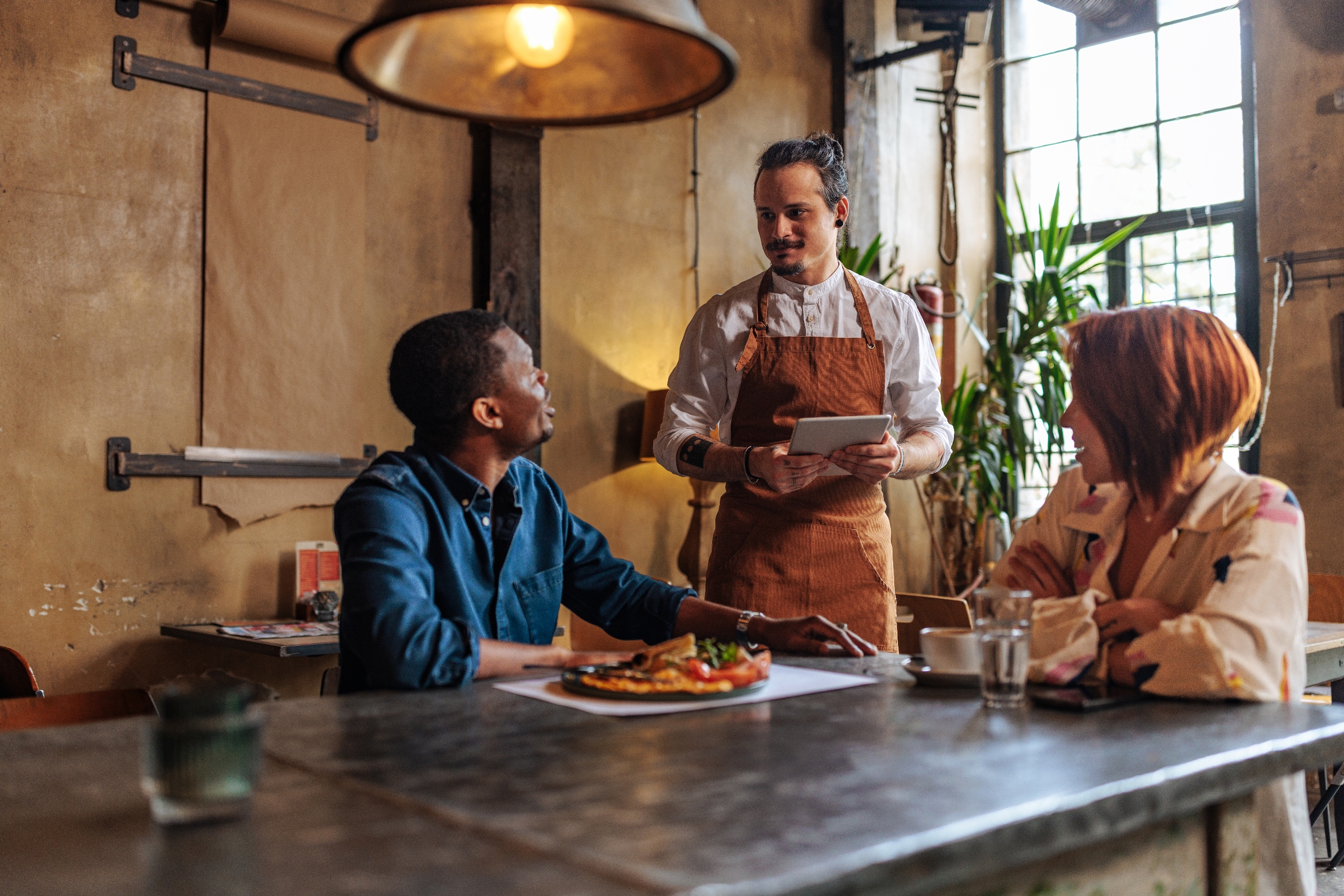 Server taking order at a cozy café, with a couple seated at a table. They are engaged and smiling, discussing their food choices
