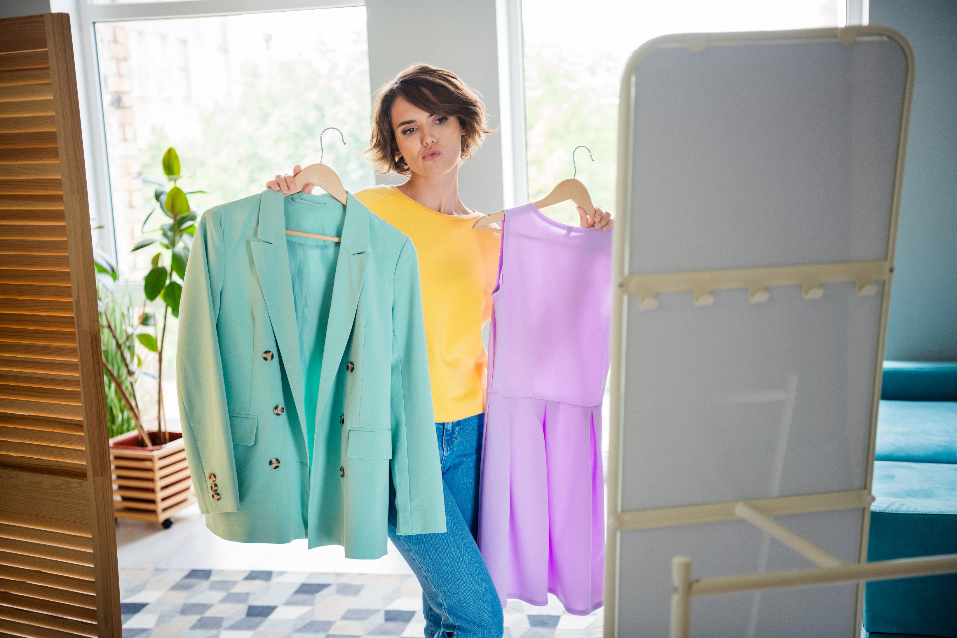 A person holds a mint blazer and a lavender dress, contemplating style choices at home with a confident expression