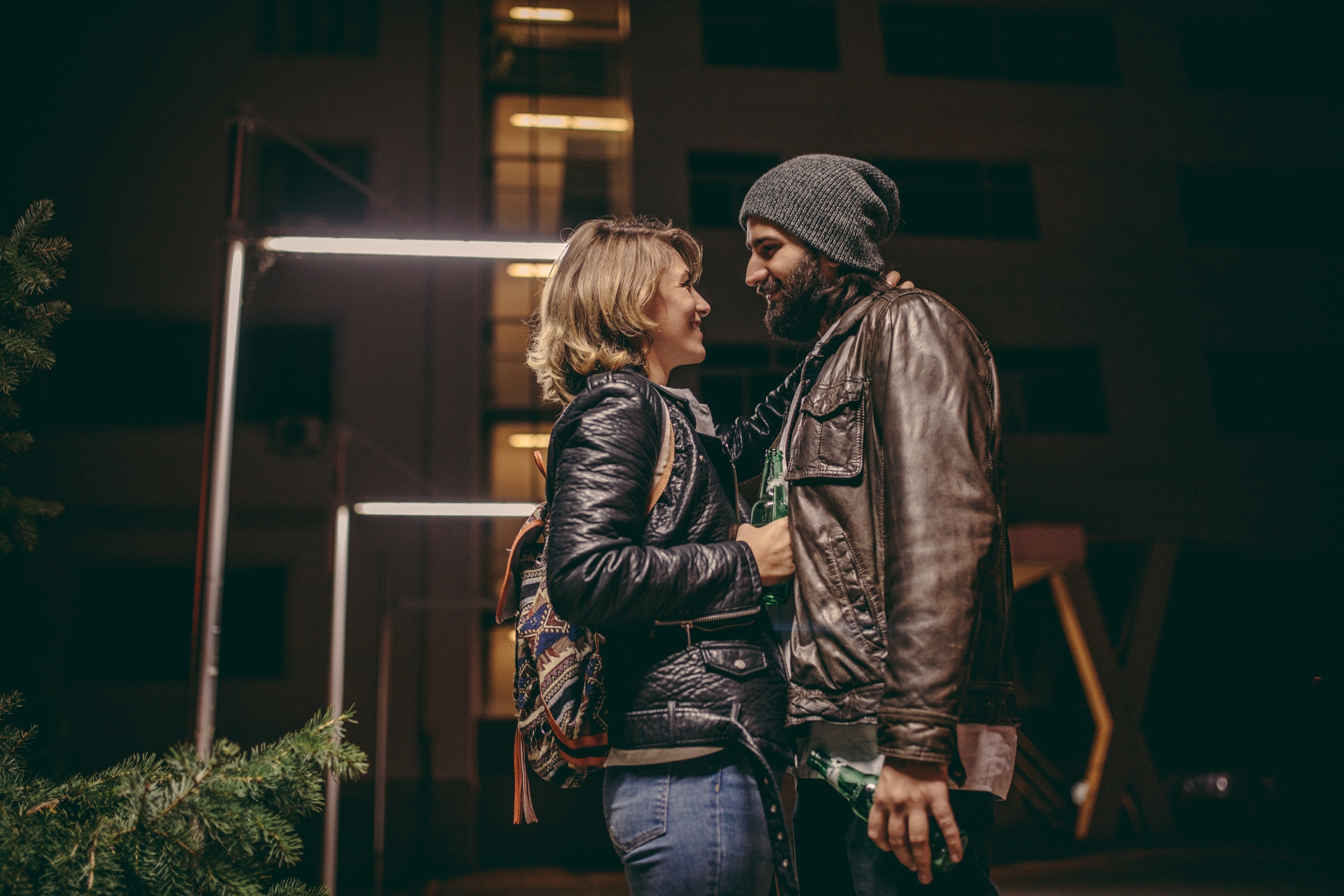 A couple wearing casual jackets and beanies shares a tender moment outside a building at night