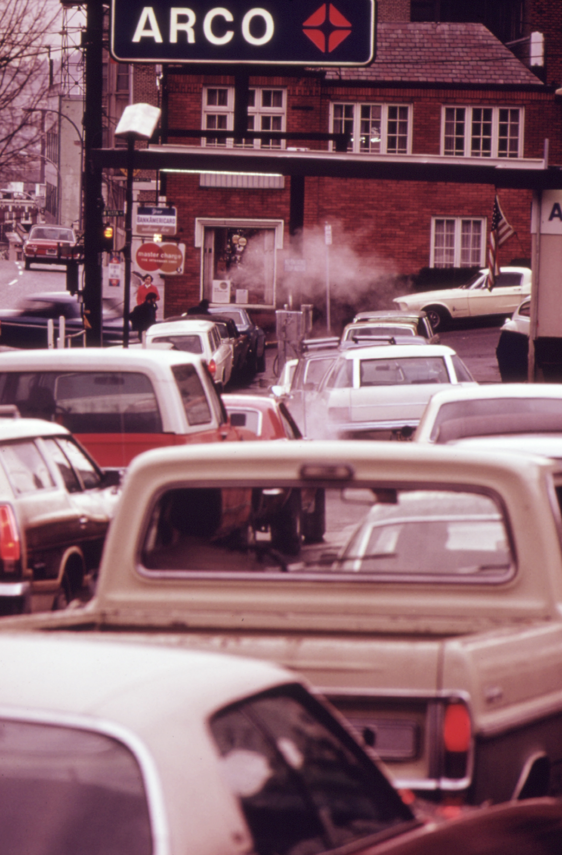 Traffic jam at a busy city intersection near a gas station. Multiple cars are waiting, with a smokestack from a chimney visible in the background