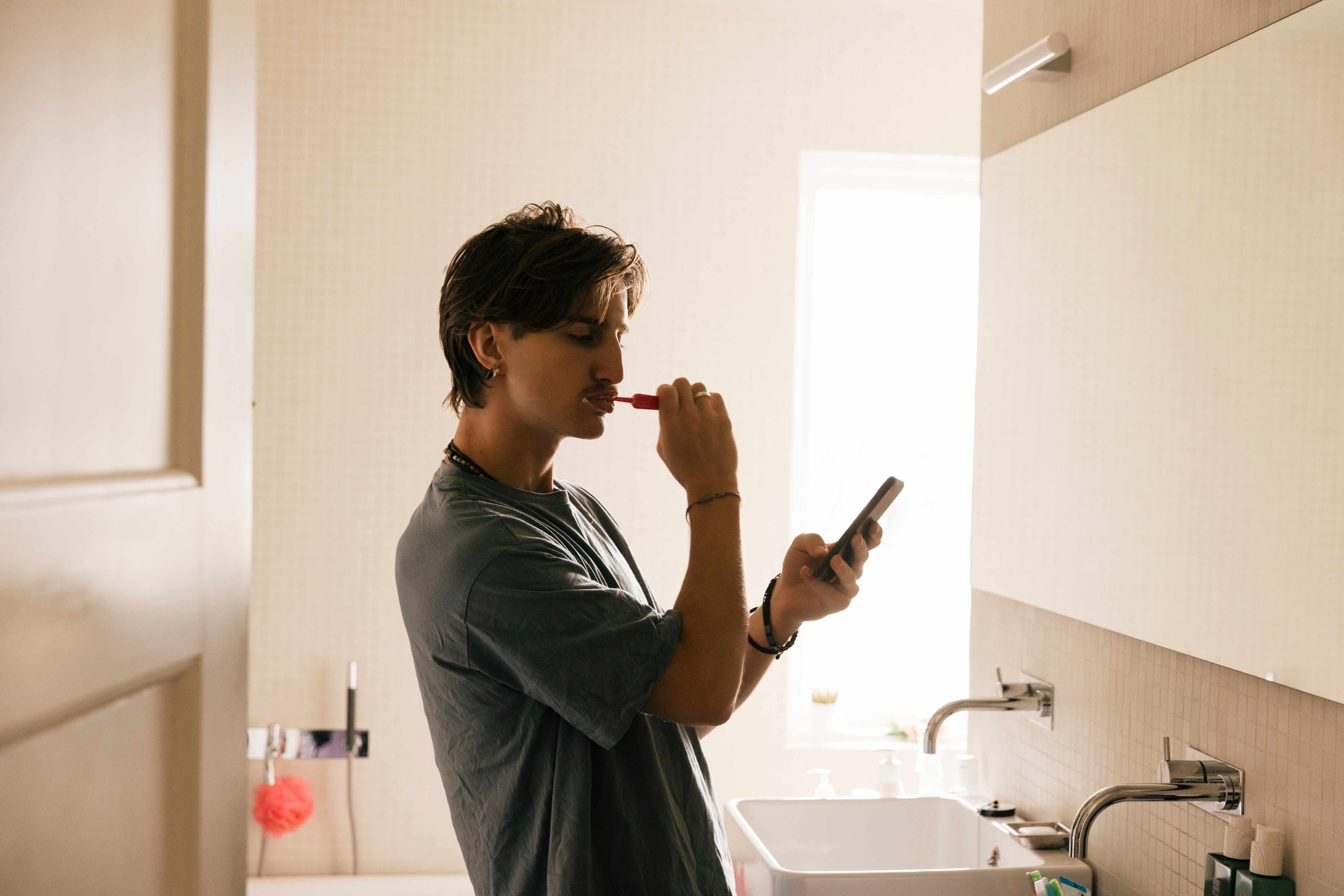 Person brushing teeth while looking at phone in a bathroom, conveying multitasking in a casual, intimate setting