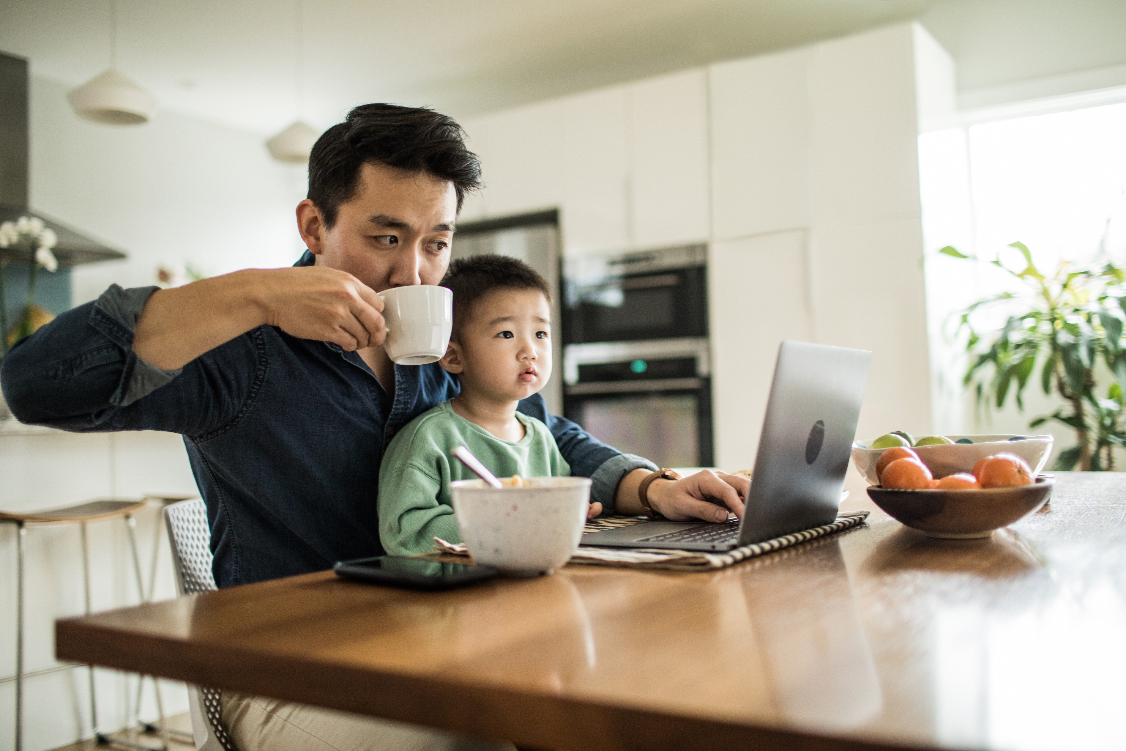 Man sipping coffee works on laptop with child sitting next to him at a kitchen table, looking at the screen. Bowl and fruit on table