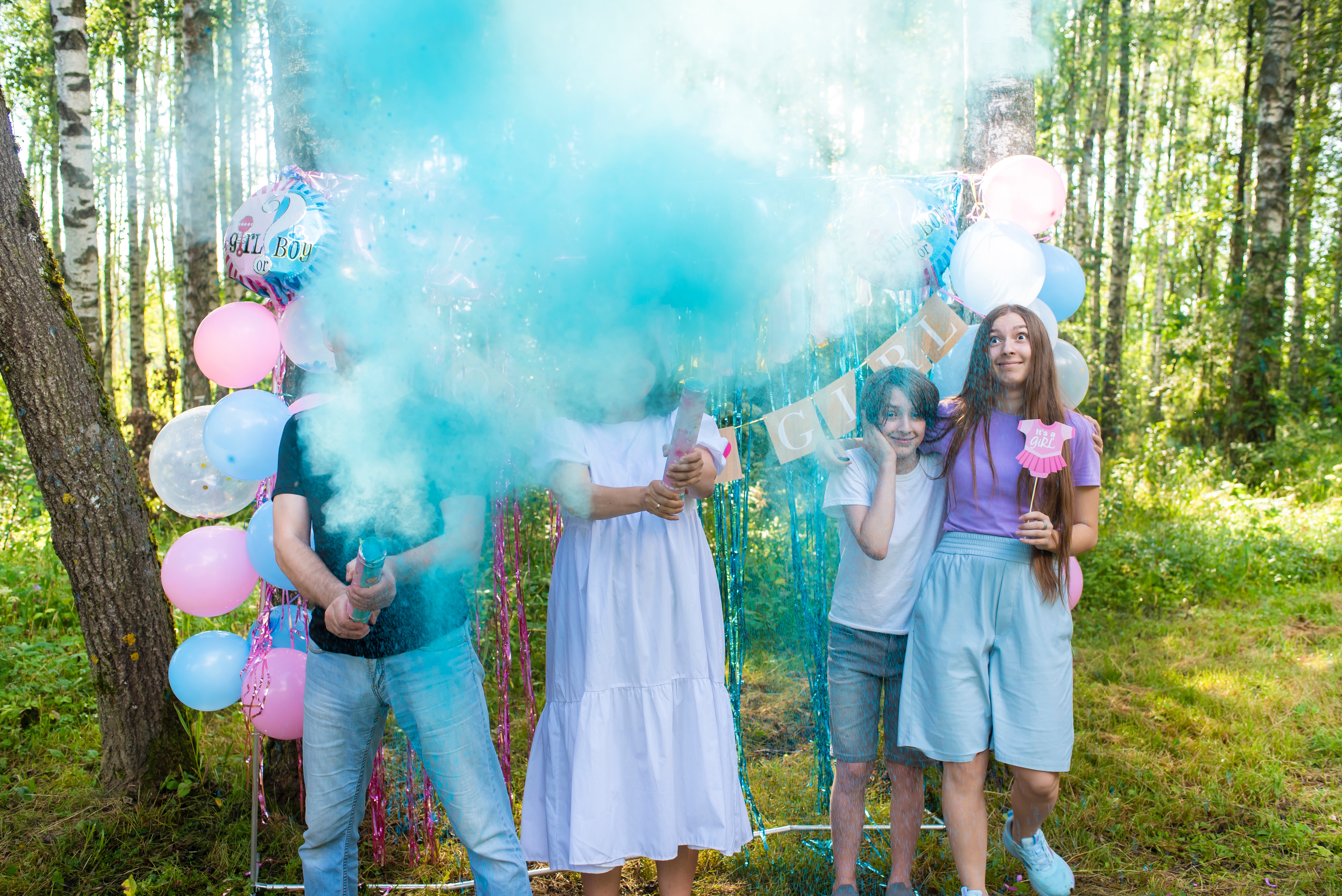 People in a forest celebrate with balloons and a blue smoke bomb, suggesting a gender reveal or celebration event