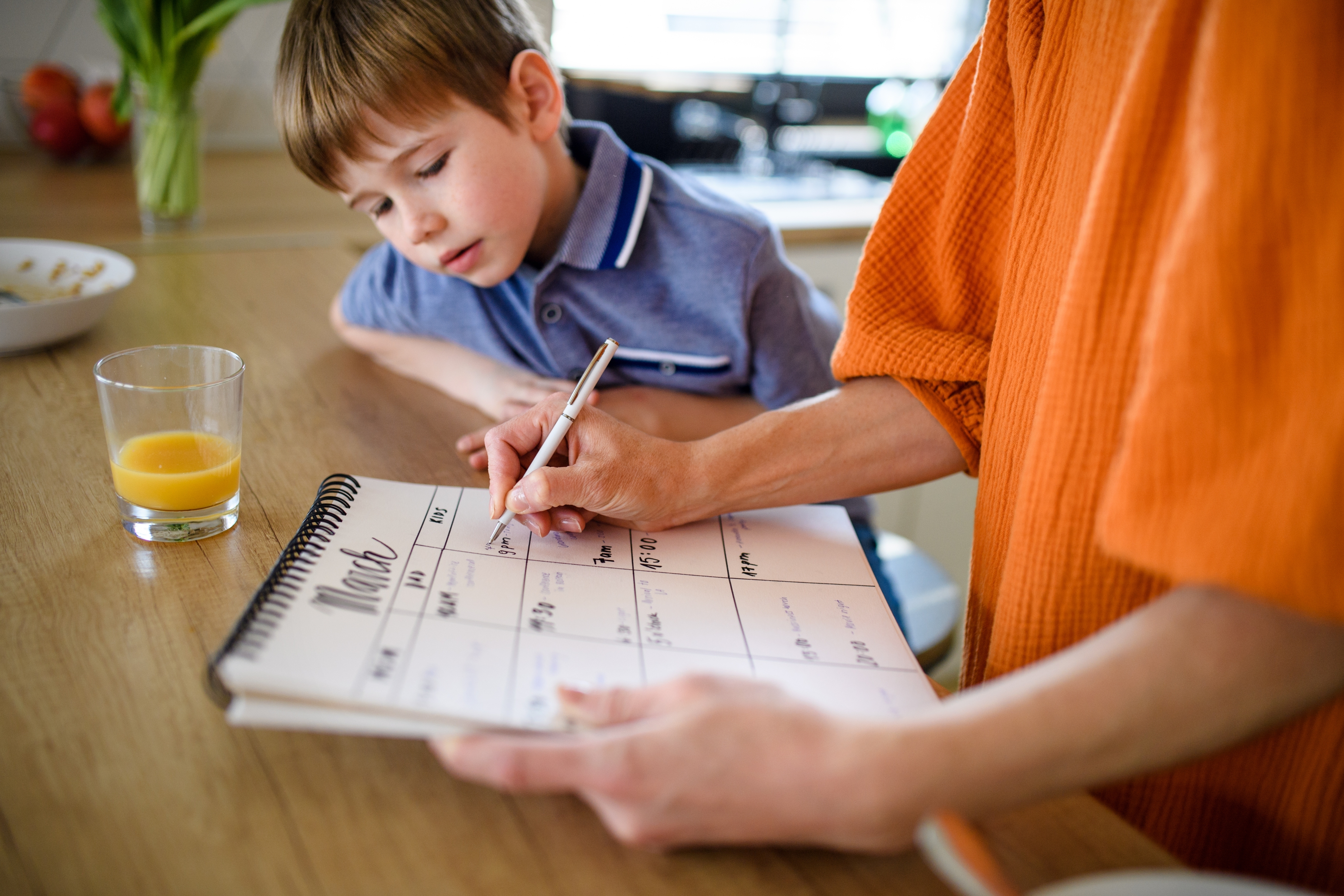 Child in polo shirt watches adult writing in a calendar at a kitchen counter. A glass of juice and a fruit bowl are nearby