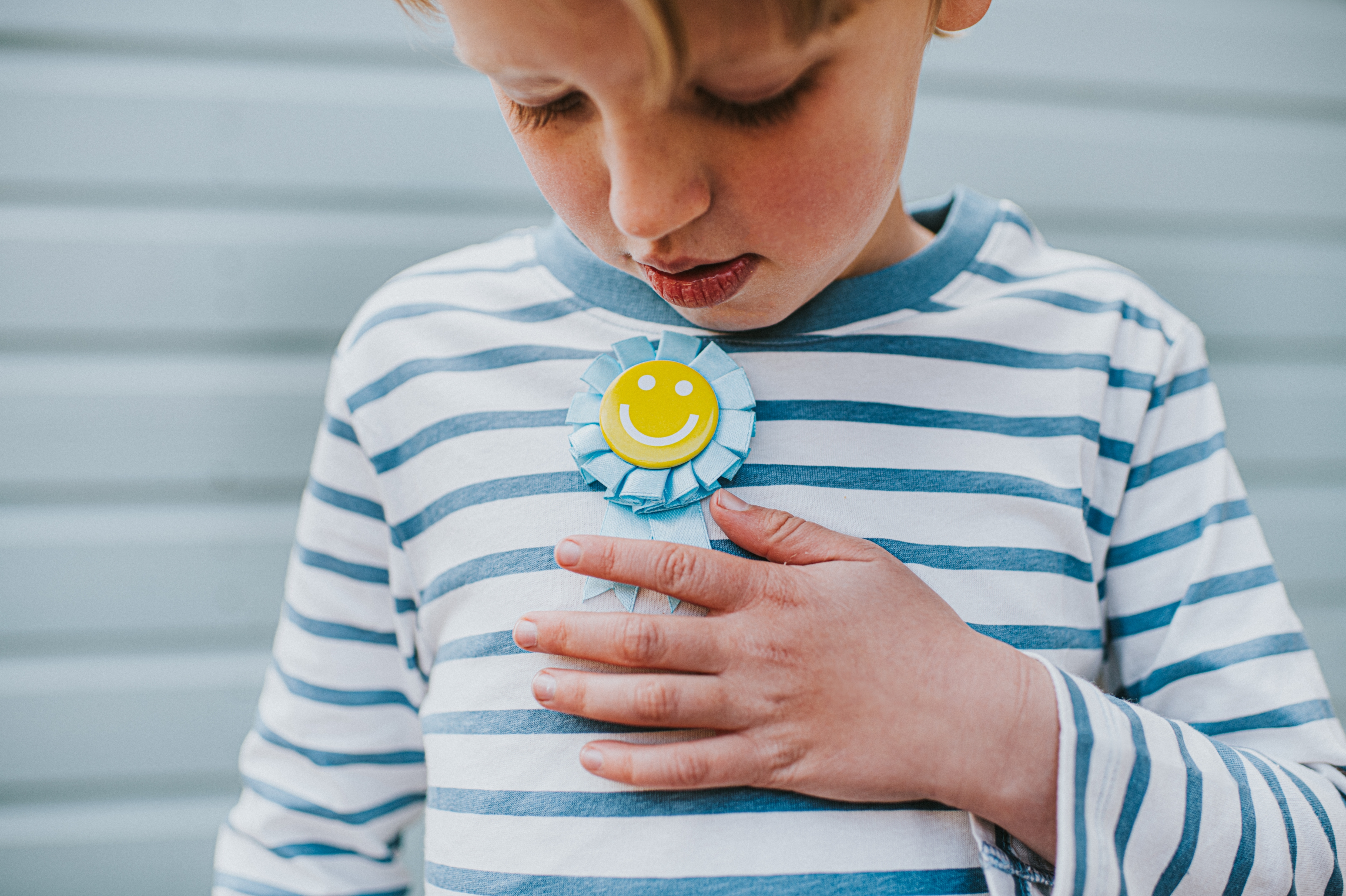 Child in striped shirt holding a smiley face badge on their chest, looking down at it thoughtfully
