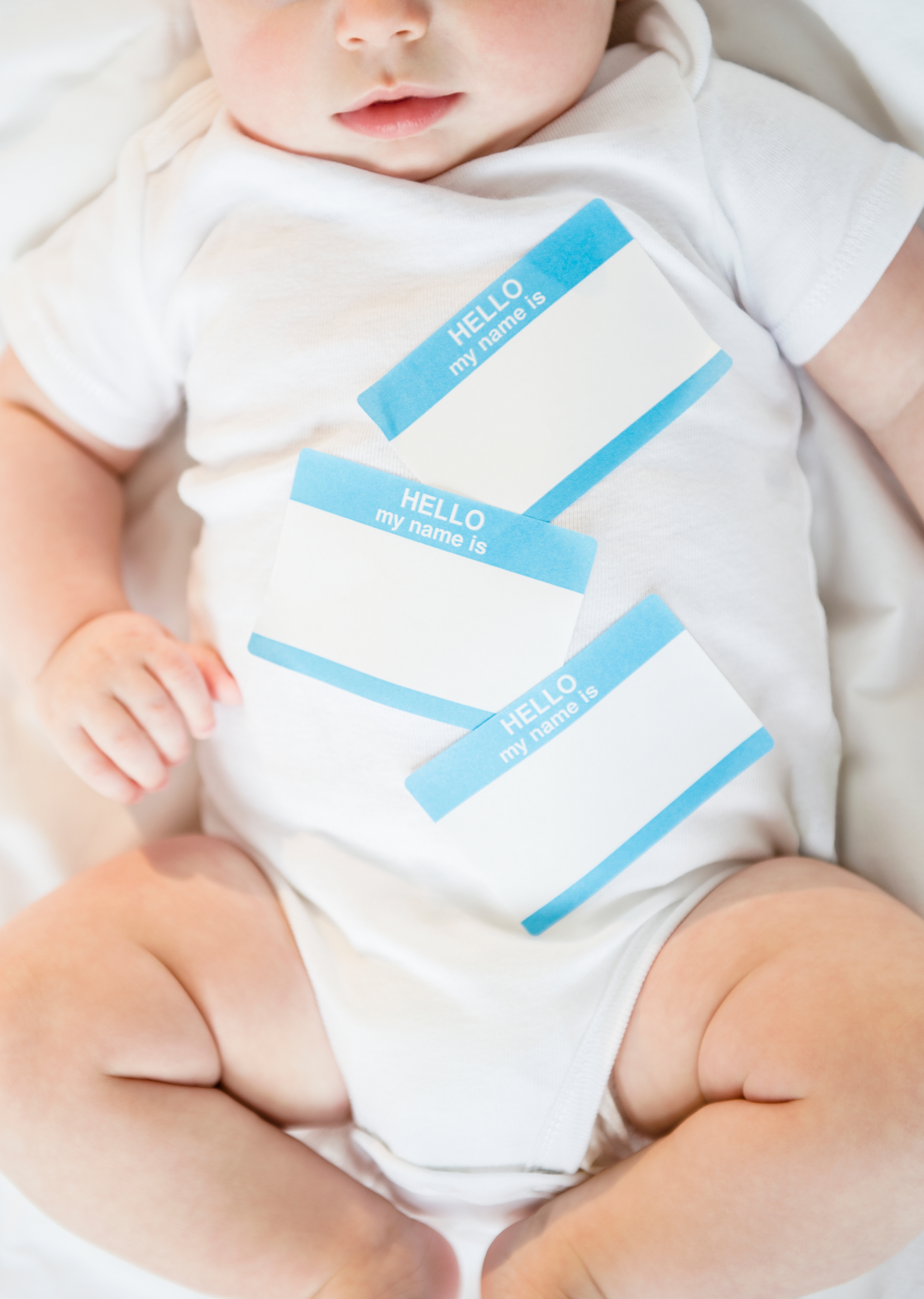 Baby lying on a white blanket wearing a onesie, with three blank &quot;Hello, my name is&quot; stickers on their chest