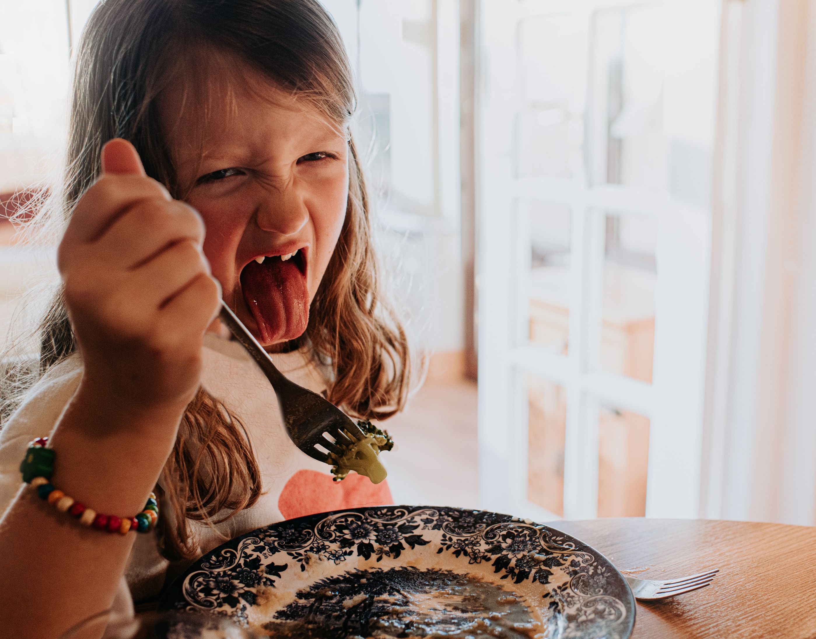 Child making a funny face while holding a fork with a piece of broccoli, sitting at a table with a decorative plate