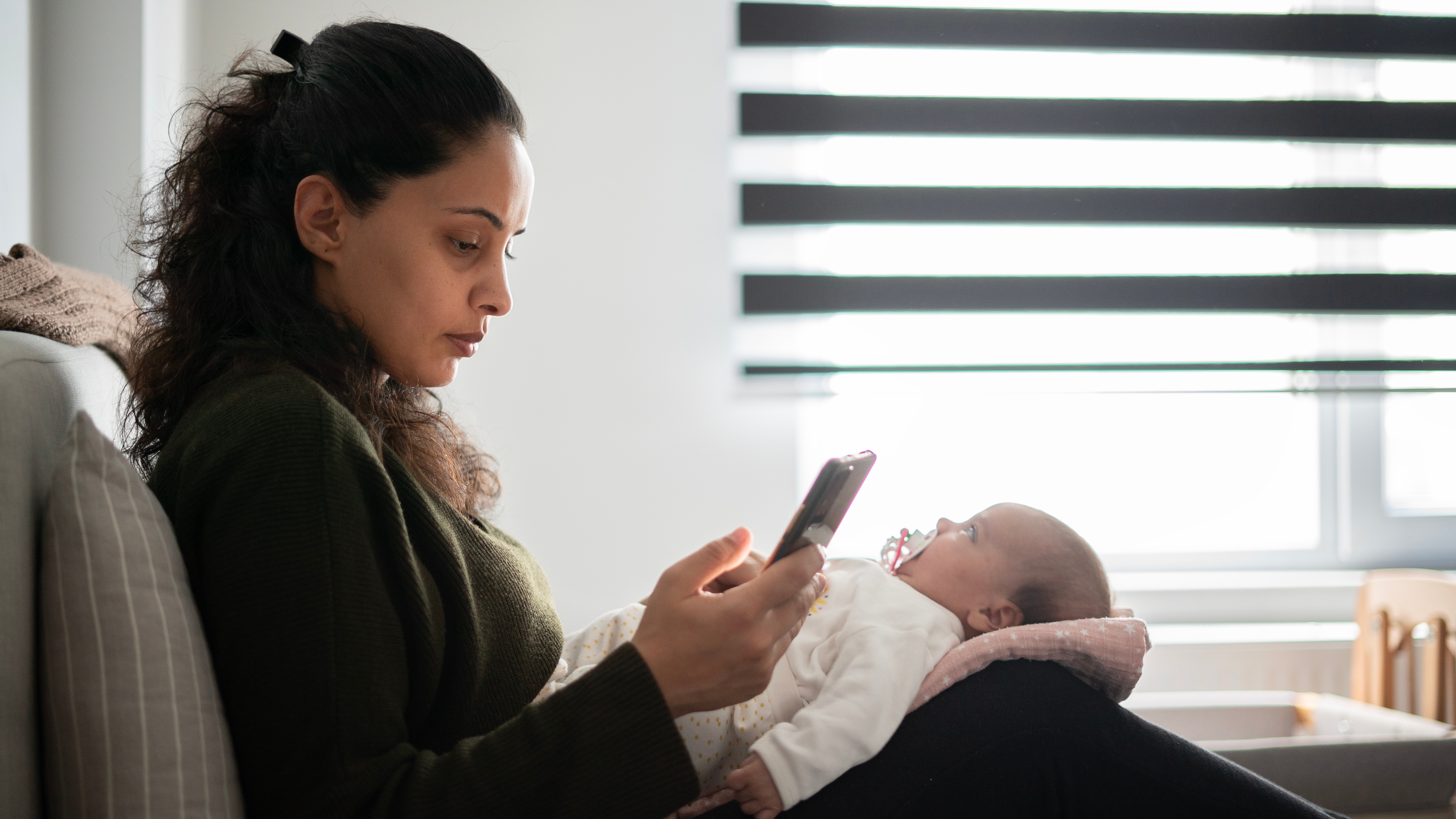 A woman sits on a sofa, looking at her phone while holding a baby on her lap, who is lying on a pillow