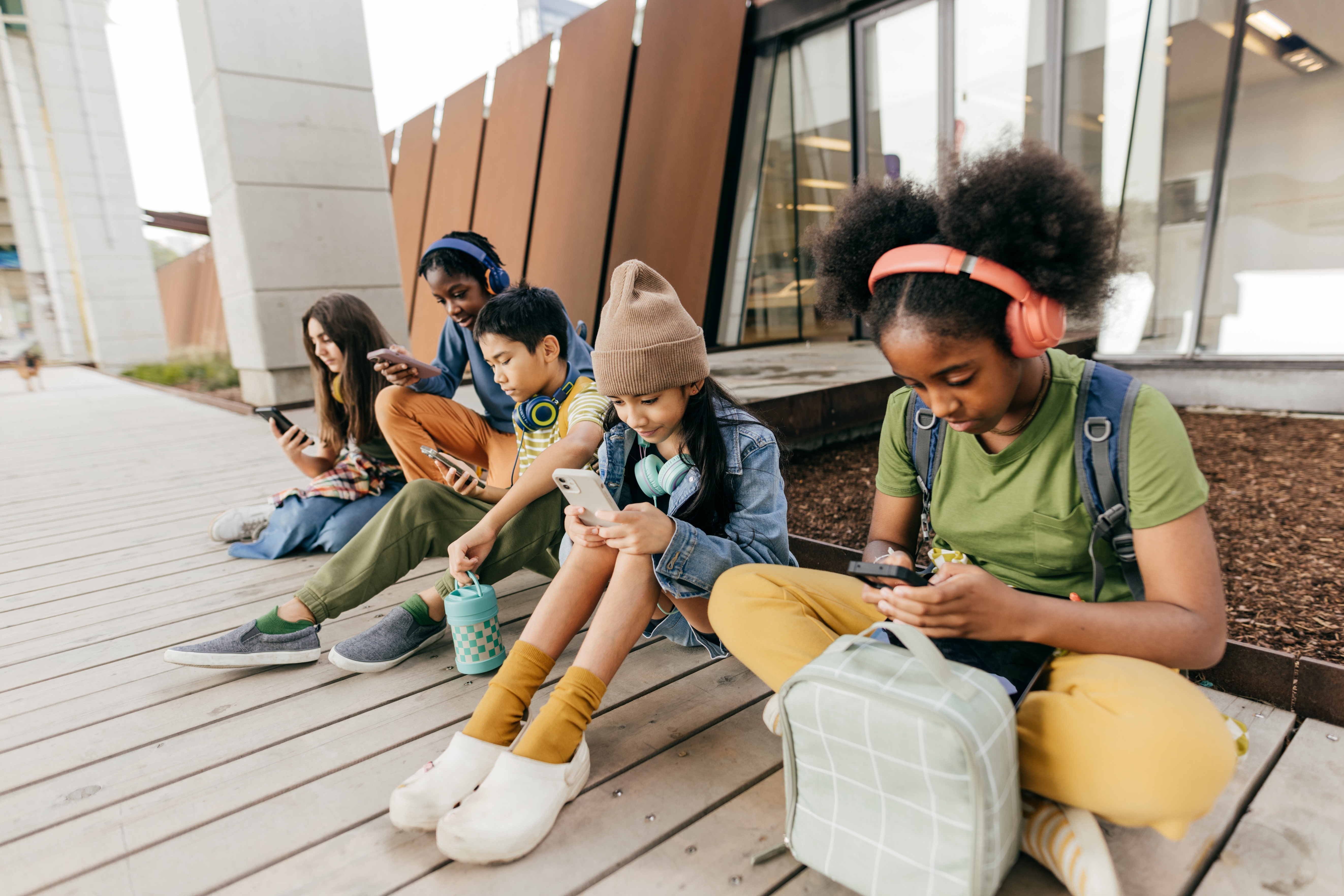 Five teens sit on a wooden deck, each using a smartphone or tablet. They wear casual clothes and headphones, engaged with their devices