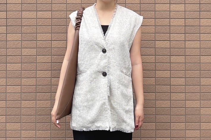 人物がノースリーブのベストを着用し、肩にバッグをかけてレンガ模様の壁の前に立っている。