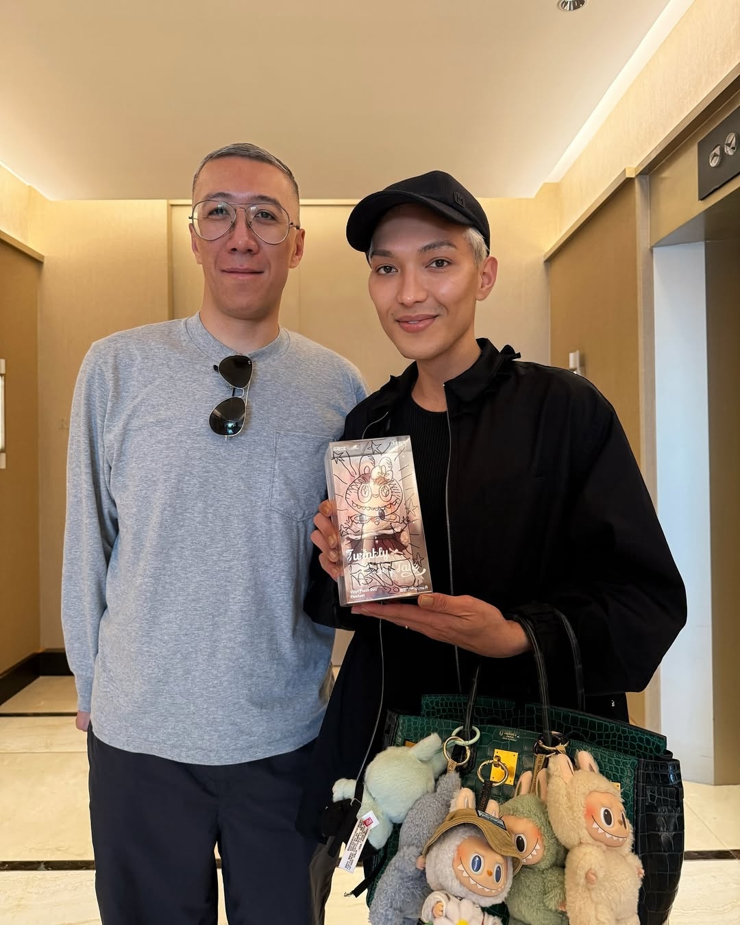 Two people pose indoors; one holds a gift and the other carries a bag adorned with stuffed toys. Both appear relaxed and casual