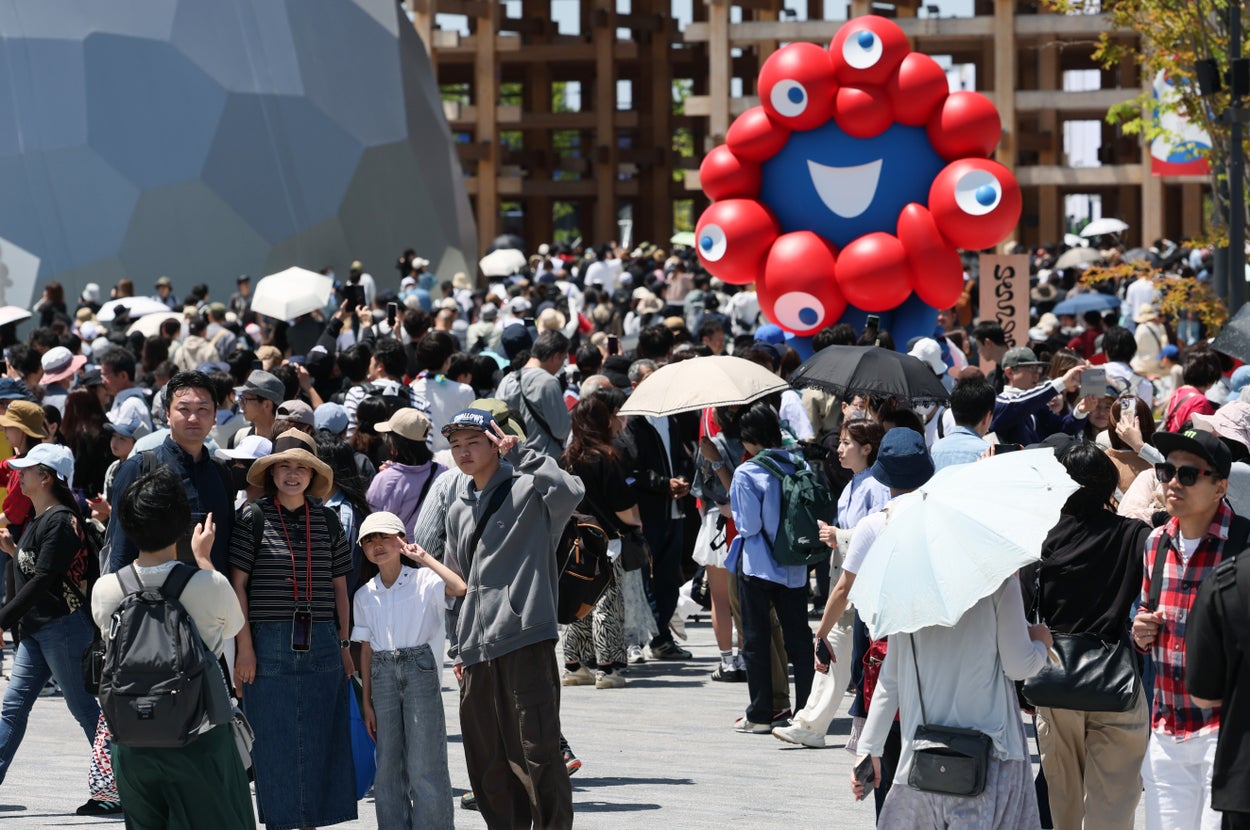 多数の人々が集まるイベント会場で、巨大な瞳付きの赤いオブジェが展示されている。多くの人が傘をさしている。