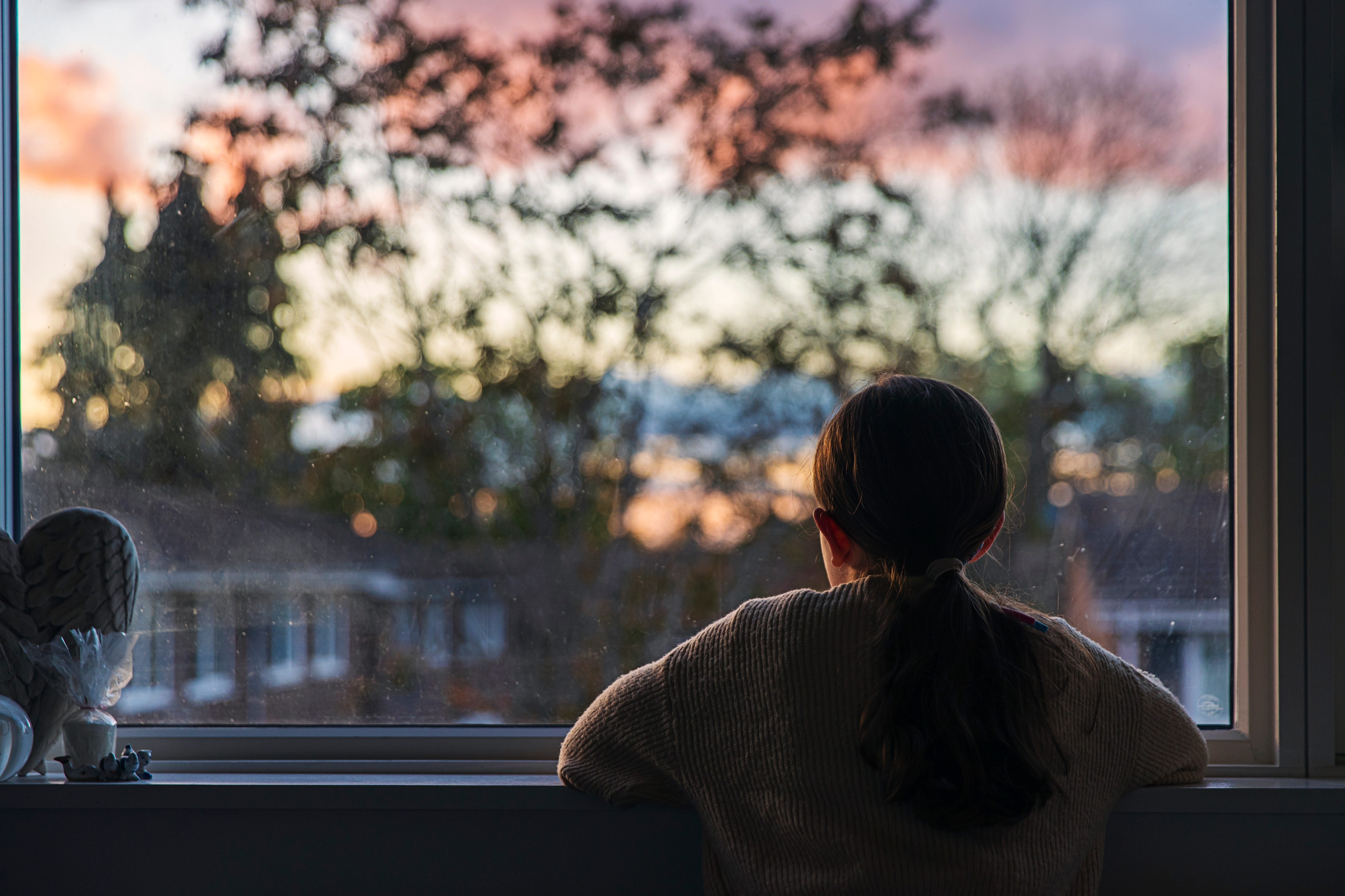 Person gazes out a window, watching a sunset beyond the silhouette of trees and houses, evoking a sense of calm reflection