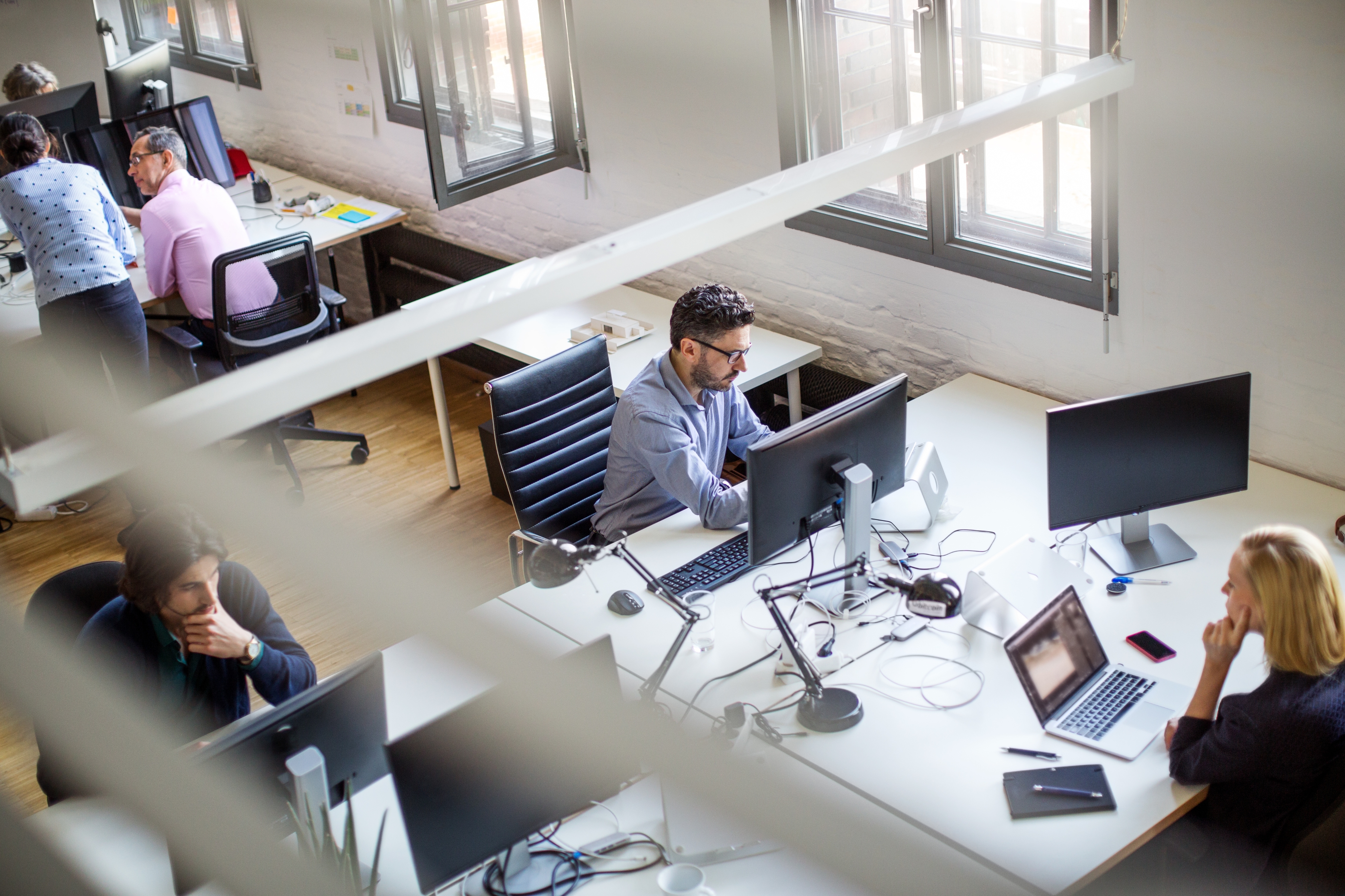 Open office space with people working at desks with computers. Natural light from large windows brightens the room