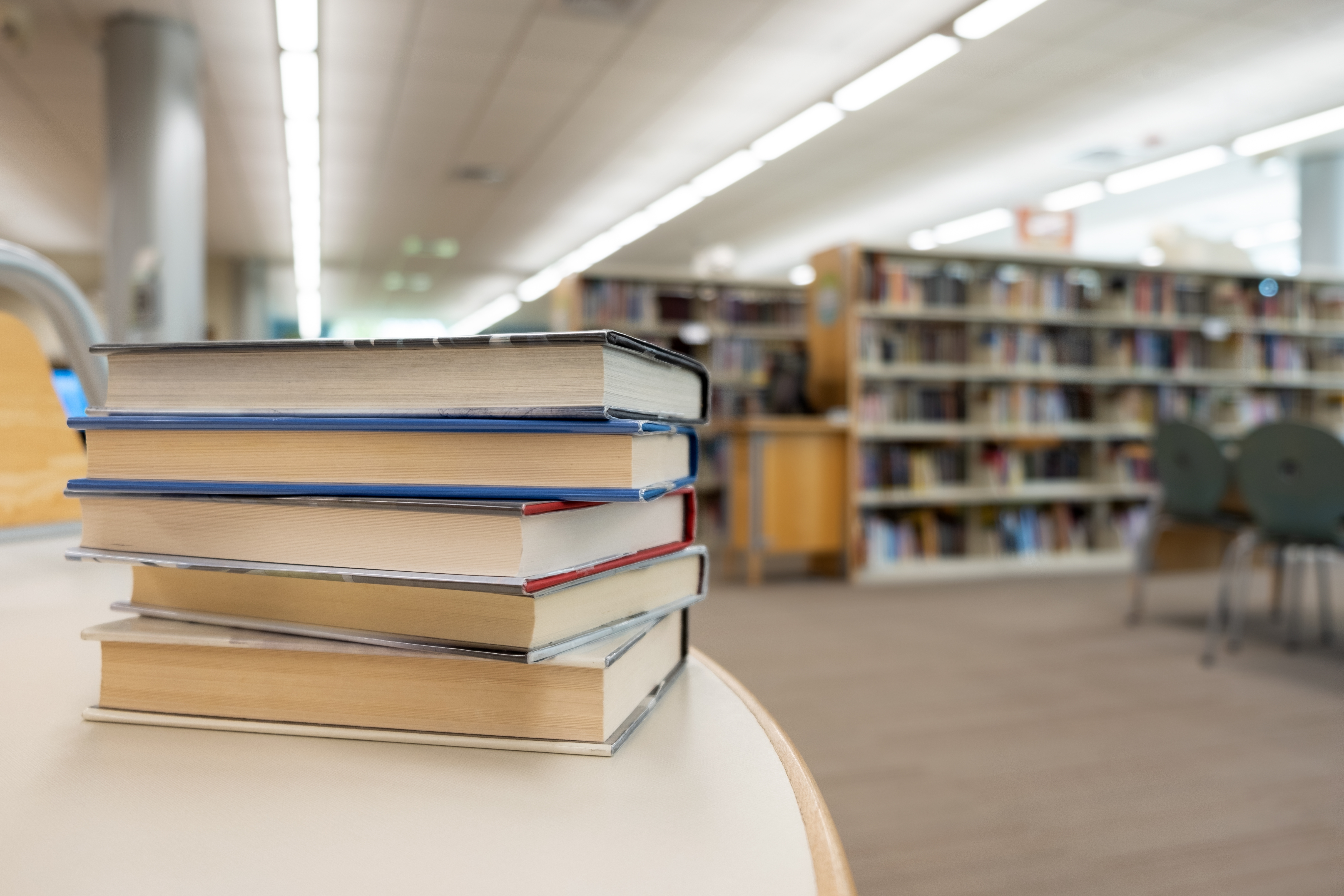 Stack of books on a table in a library, with shelves full of more books in the background
