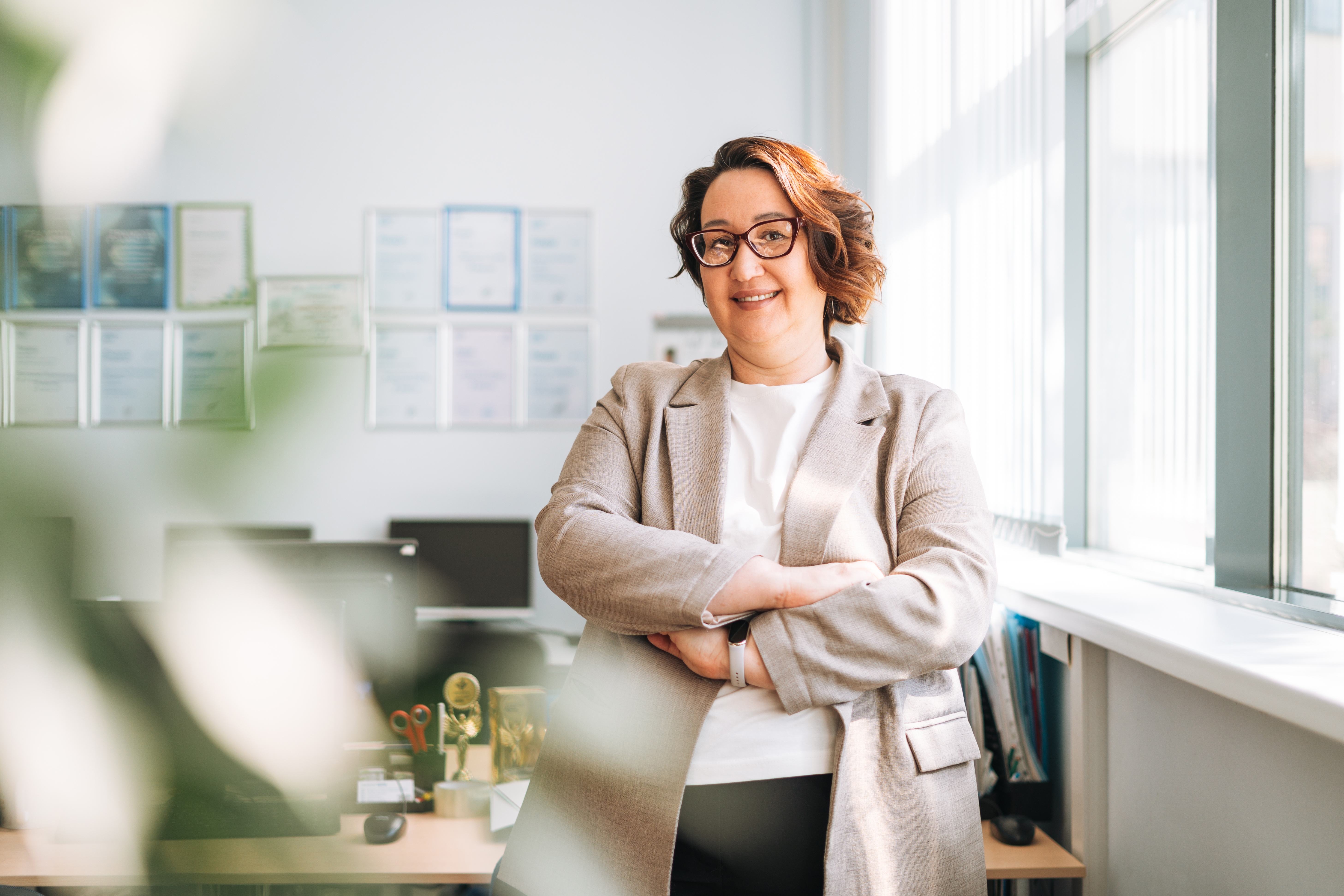Person in a professional setting, wearing glasses and a blazer, smiling with arms crossed near an office window