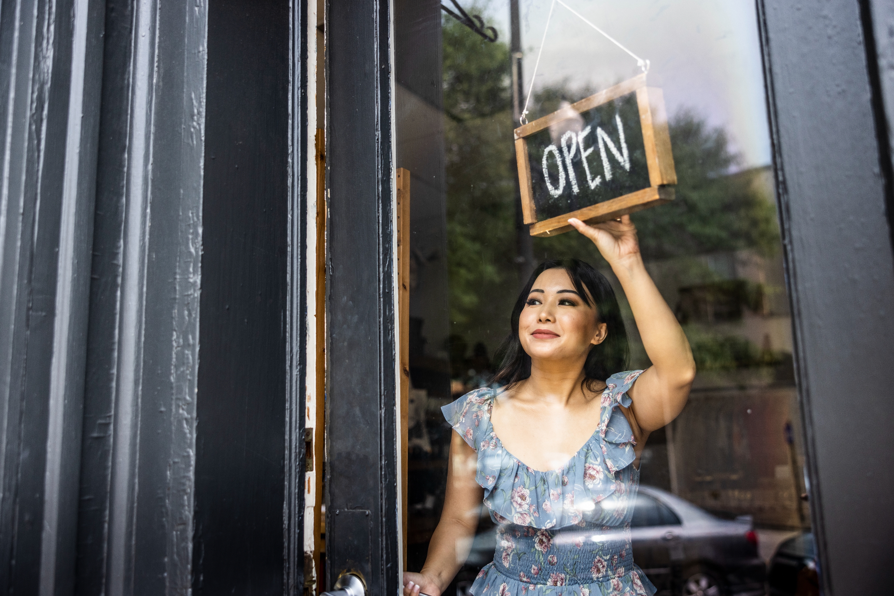 Person in a floral dress flips an "Open" sign on a glass door of a storefront