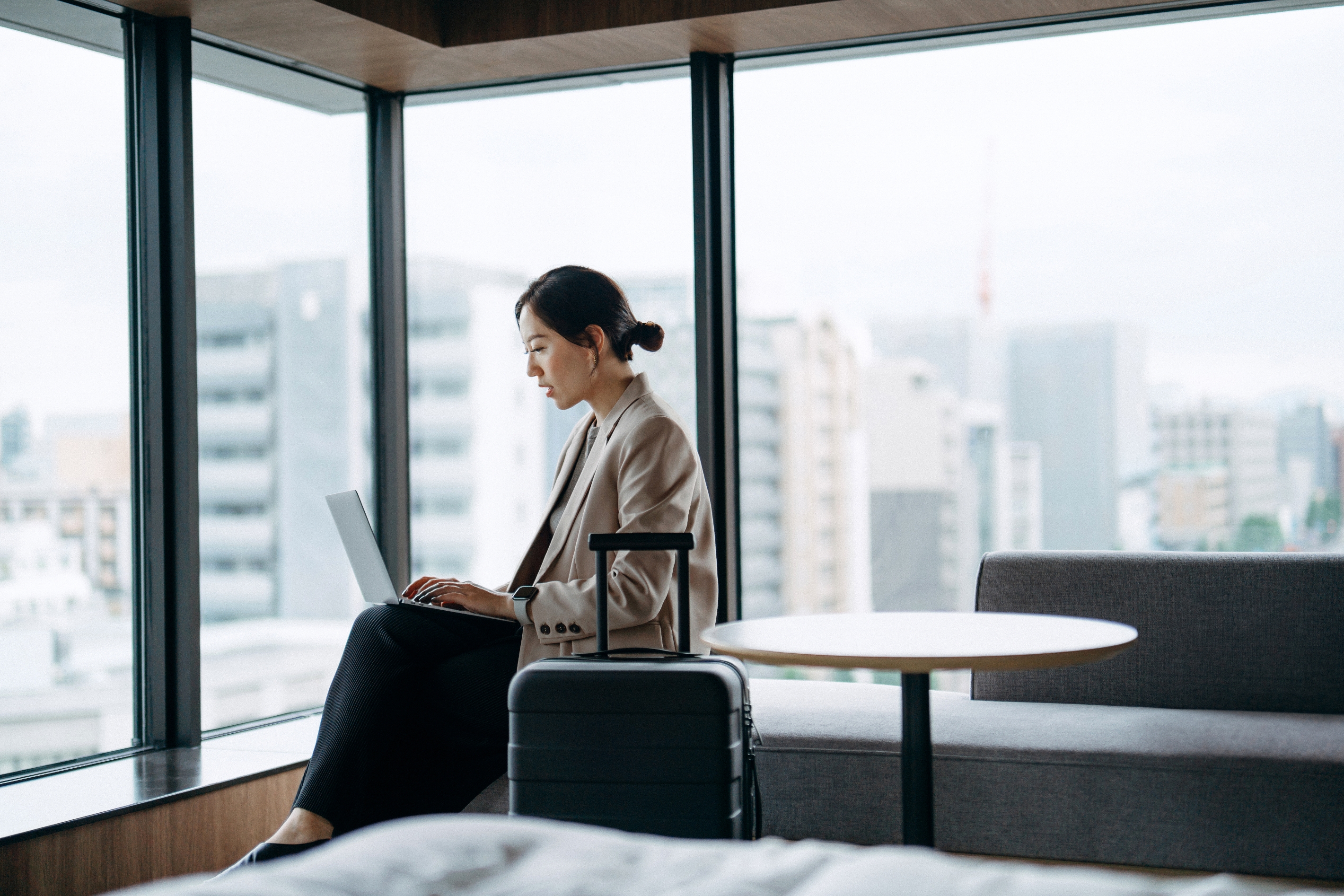 Person sitting by a window, working on a laptop, next to a suitcase in a modern, urban setting