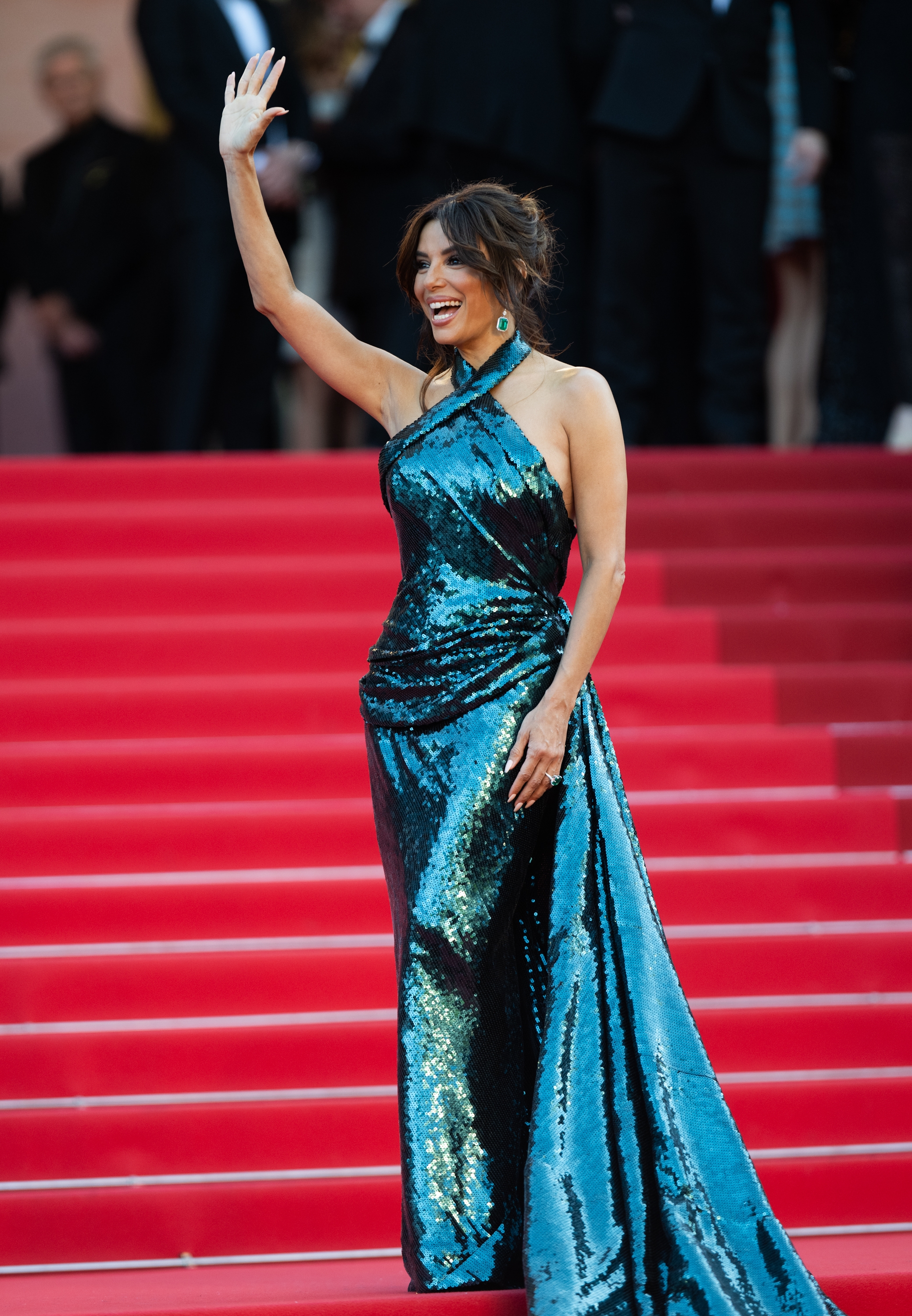 Eva Longoria in a shimmering gown waves on a red carpet, smiling amidst event attendees