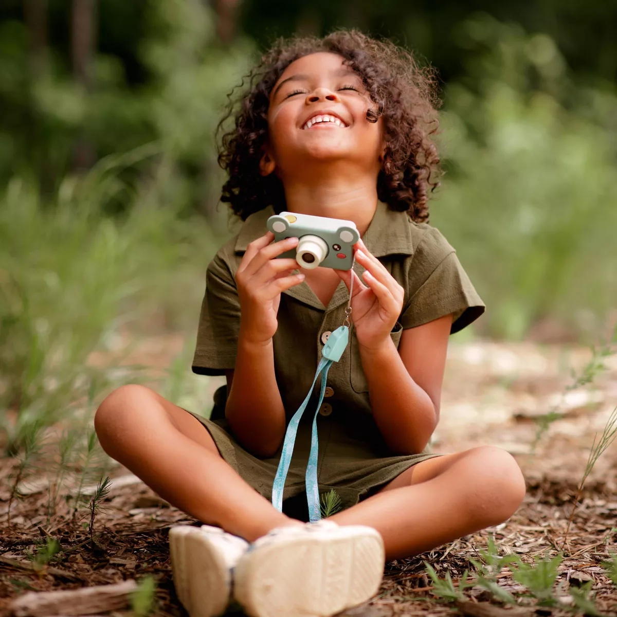 Child joyfully holding a toy camera, sitting cross-legged outdoors in a casual outfit, smiling upwards