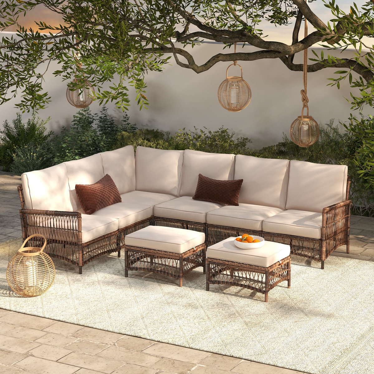 Outdoor wicker sectional with cushions, a matching ottoman, and a table with fruit, set under a tree with hanging lanterns on a patio