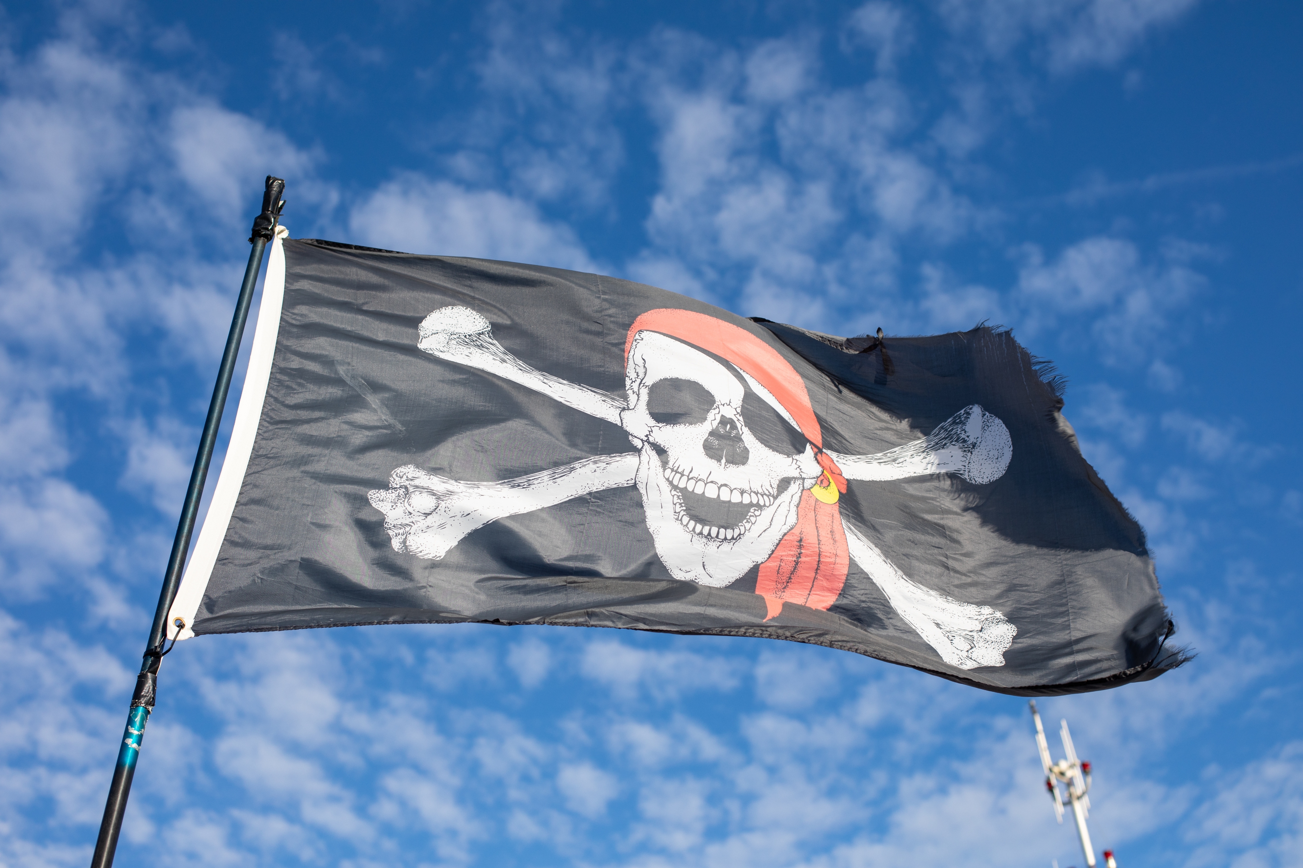 Pirate flag with skull and crossbones, wearing an eyepatch and red bandana, waves against a blue, cloud-dotted sky