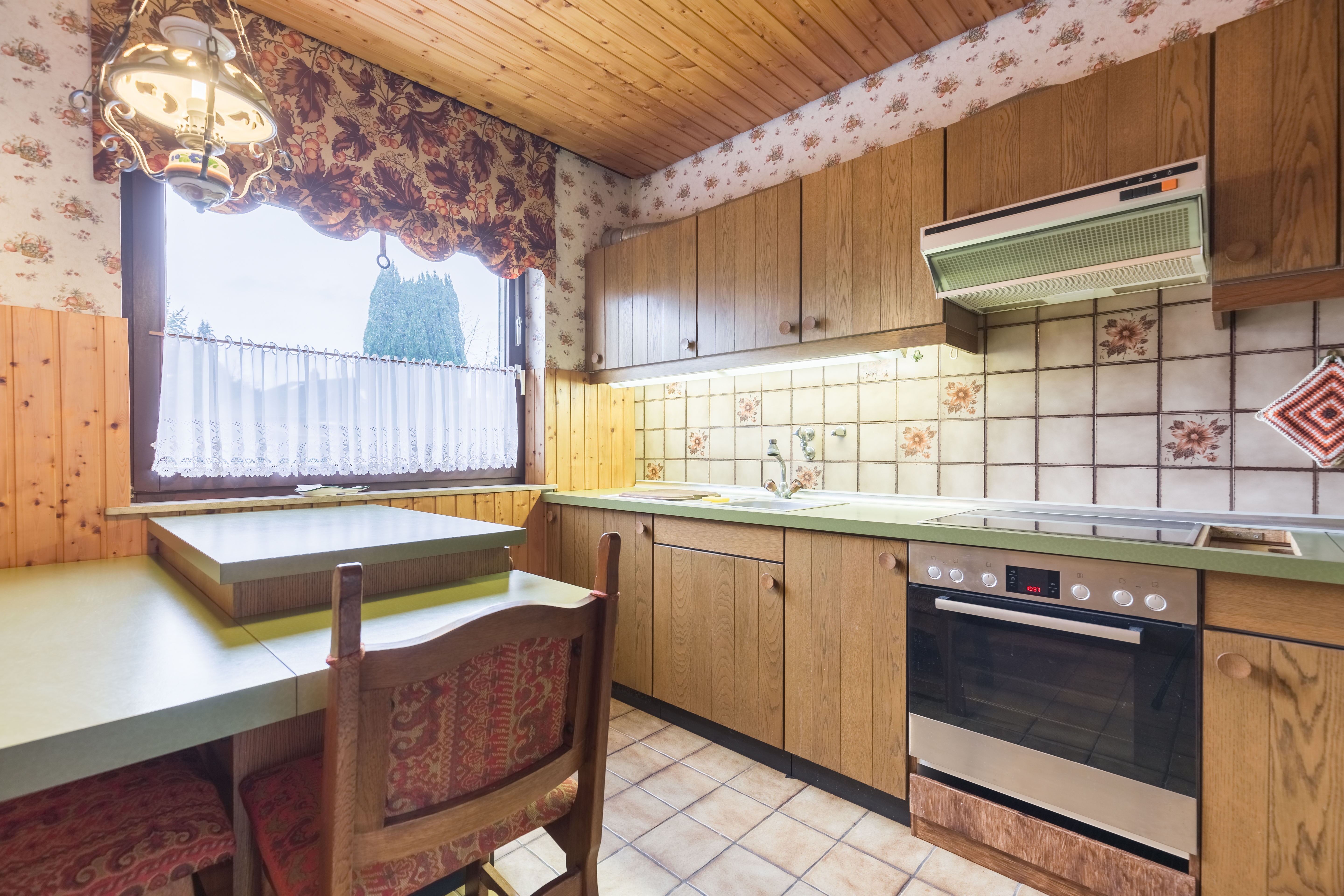 Vintage kitchen with wood cabinets, floral wallpaper, tiled backsplash, table with chairs, and modern oven under vent hood. Cozy and retro style