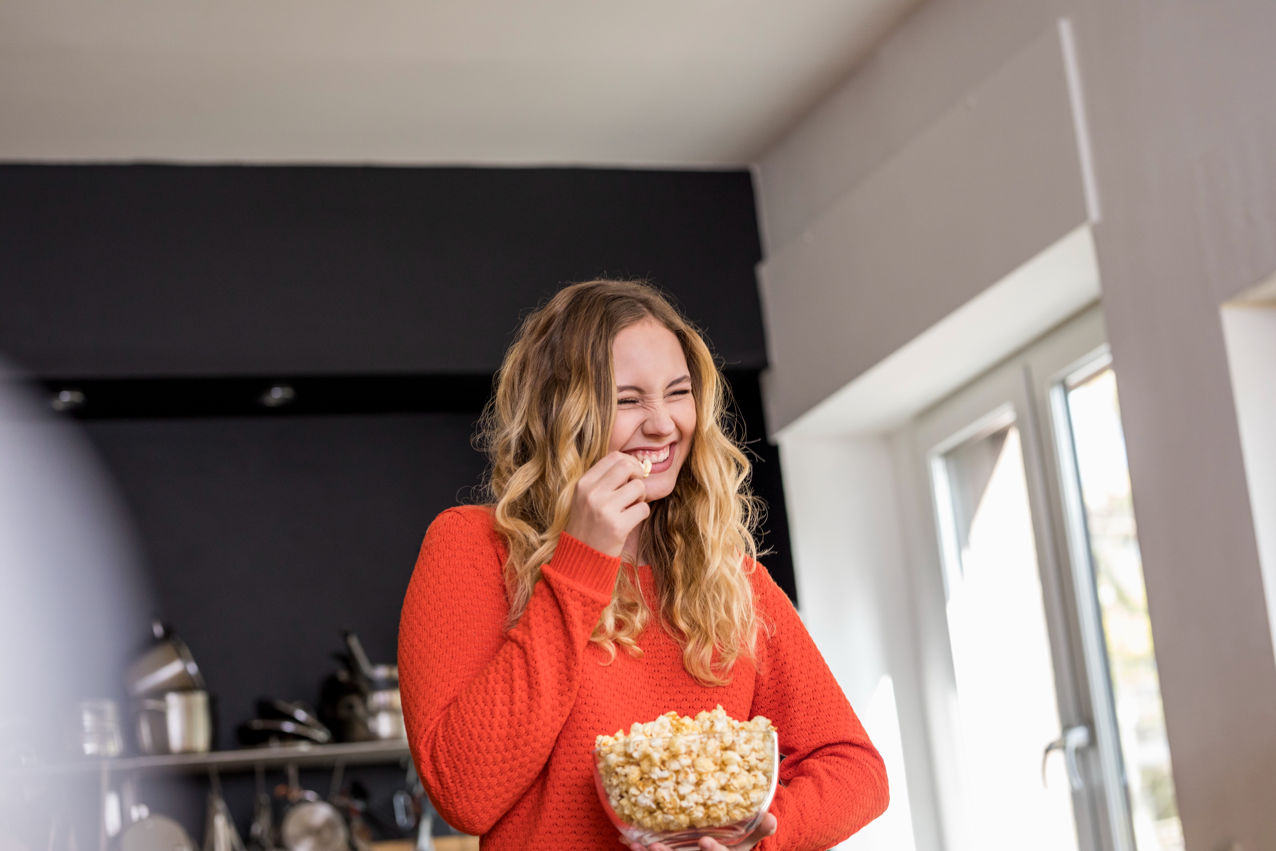A person smiling, holding a bowl of popcorn, standing in a kitchen lit by natural light