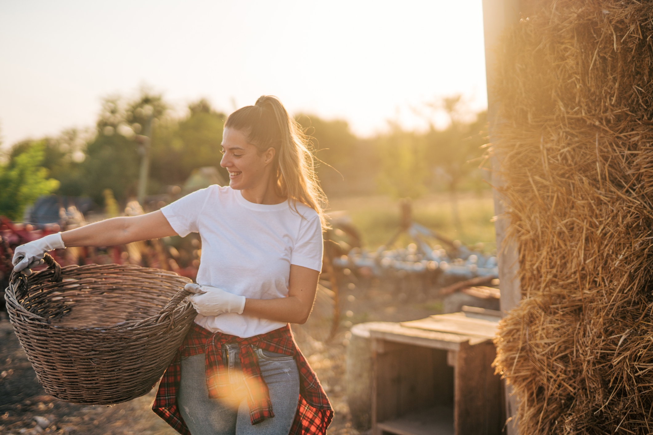 Woman smiling while holding a basket in a sunlit outdoor setting, wearing casual attire with a plaid shirt tied around her waist