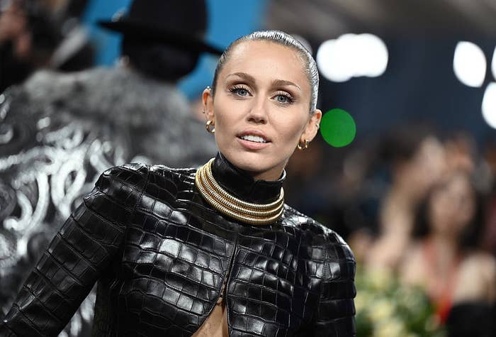 Person successful a achromatic textured outfit with aggregate golden necklaces, smiling astatine an event. Blurred radical and lights successful the background