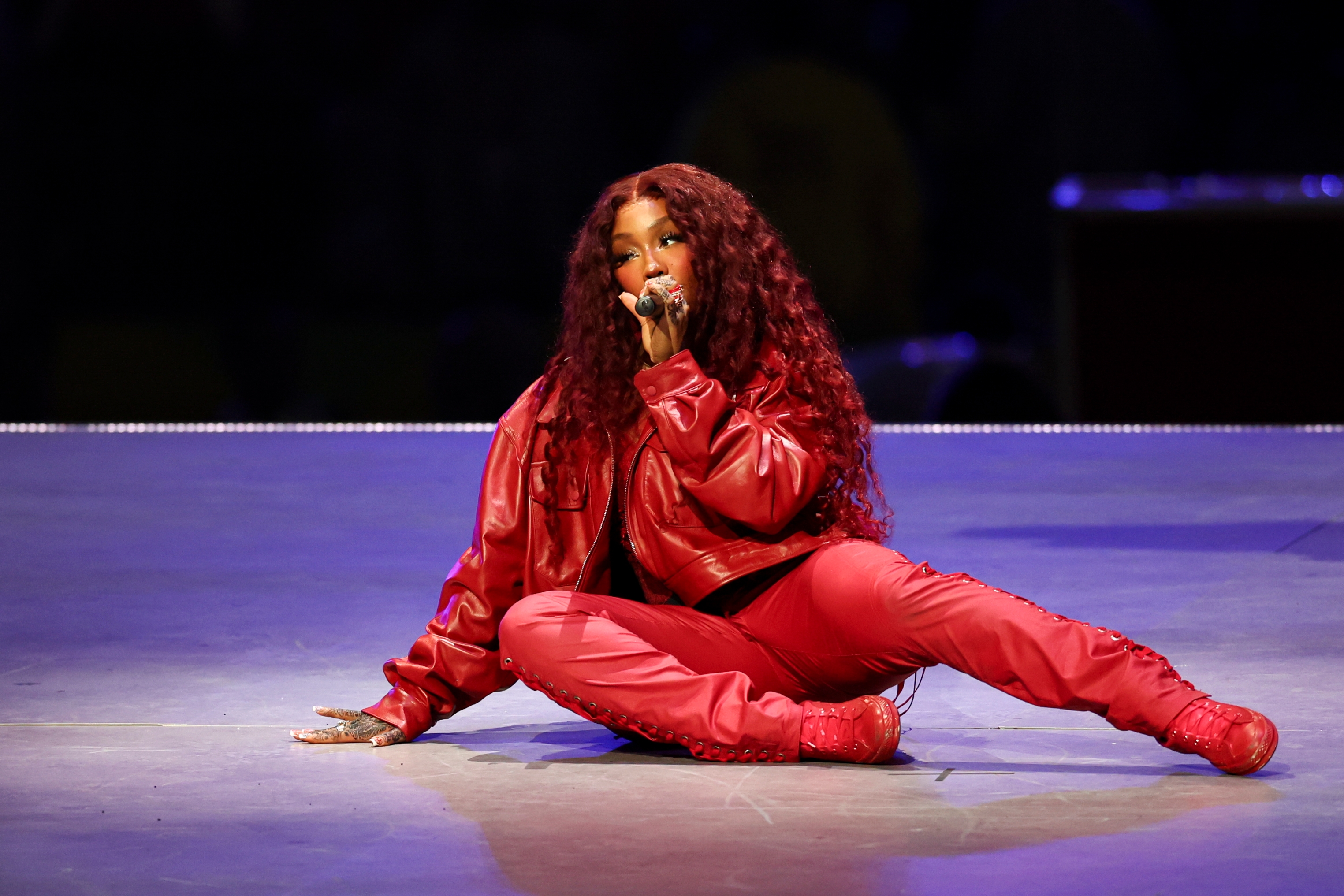 A woman performer sits onstage singing, wearing a red outfit with a jacket and fitted pants, styled with long hair