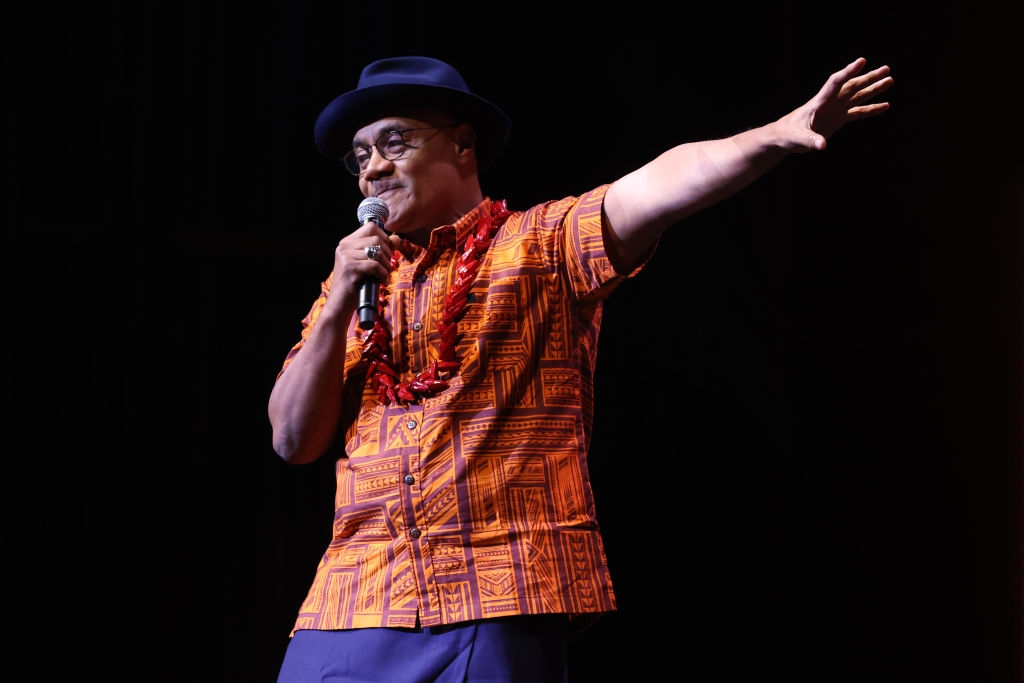 Person on stage in printed shirt, wearing a hat and lei, holding a microphone, gesturing with one arm extended