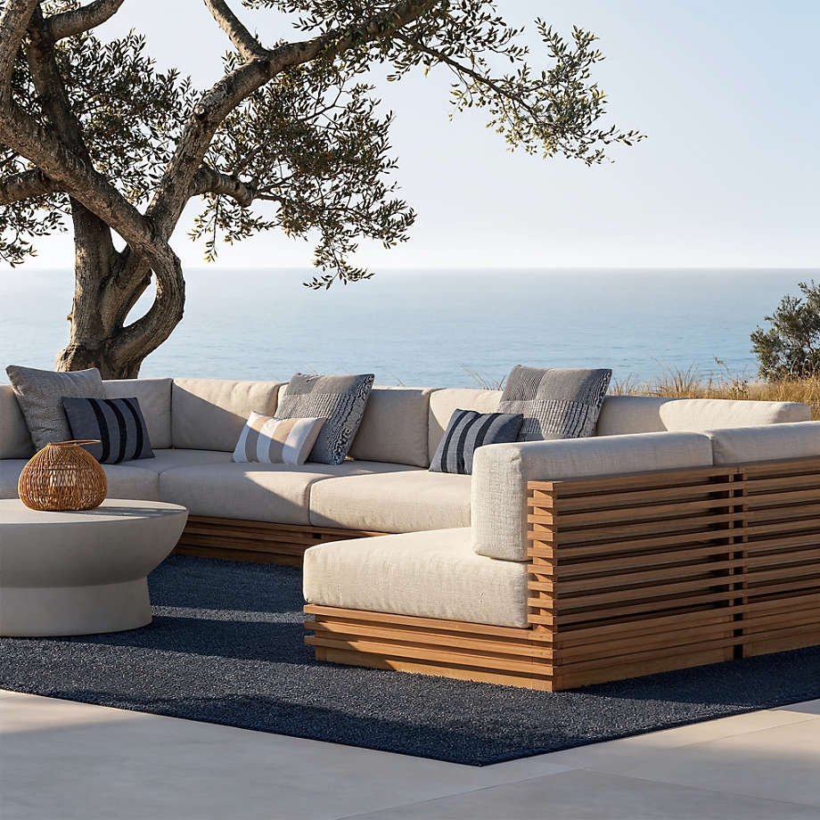 Outdoor patio with modern sectional sofa, wooden accents, and a view of the ocean; a round coffee table is placed on a textured rug