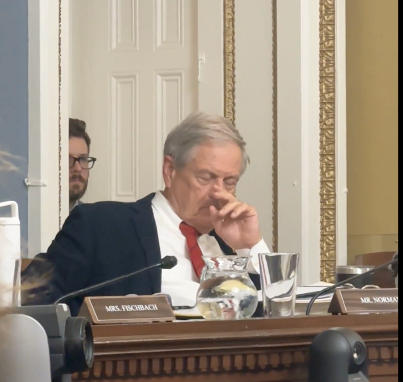 An older man in a suit and red tie sits at a governmental meeting, appearing deep in thought, with nameplates visible on the desk