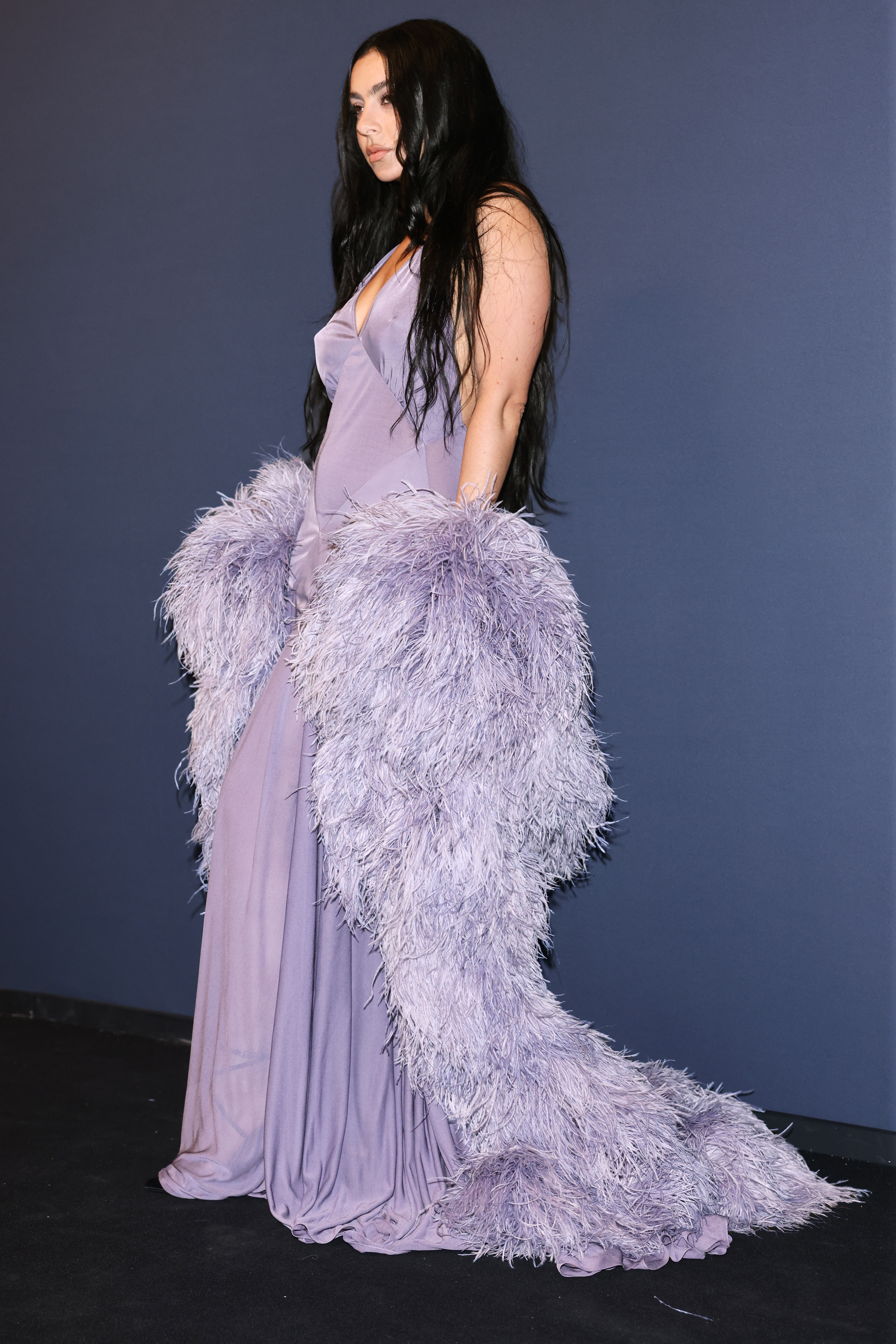 Charli XCX in a flowing gown with a feathered train, posing on a red carpet