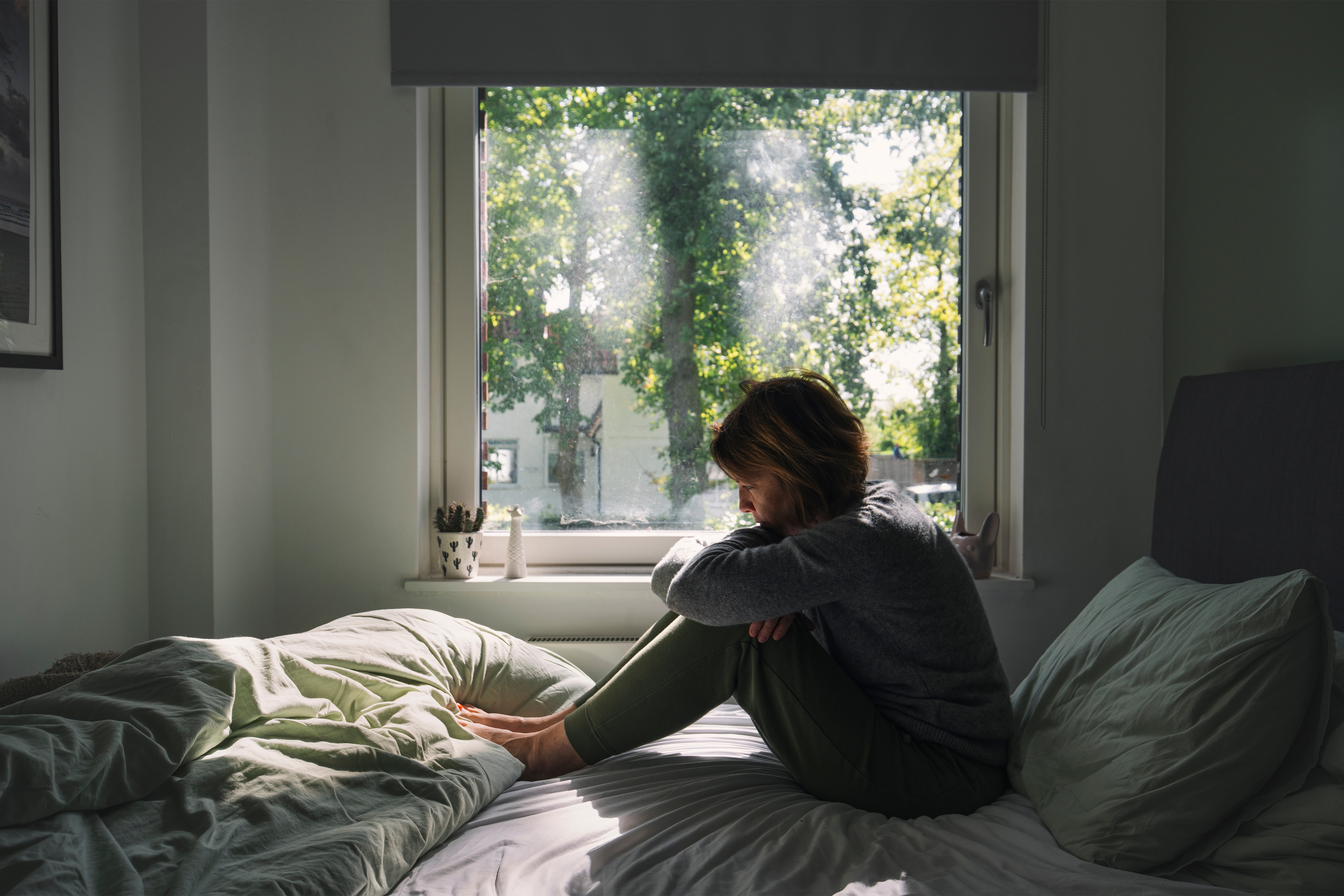 Person sitting on a bed in a sunlit room, looking outside through a window. The scene feels contemplative and peaceful