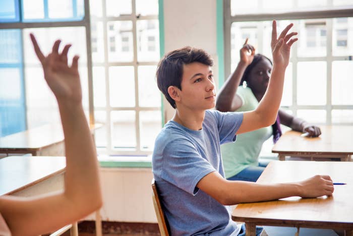 Students in a classroom raising hands to participate, seated at desks by large windows, focused on the teacher